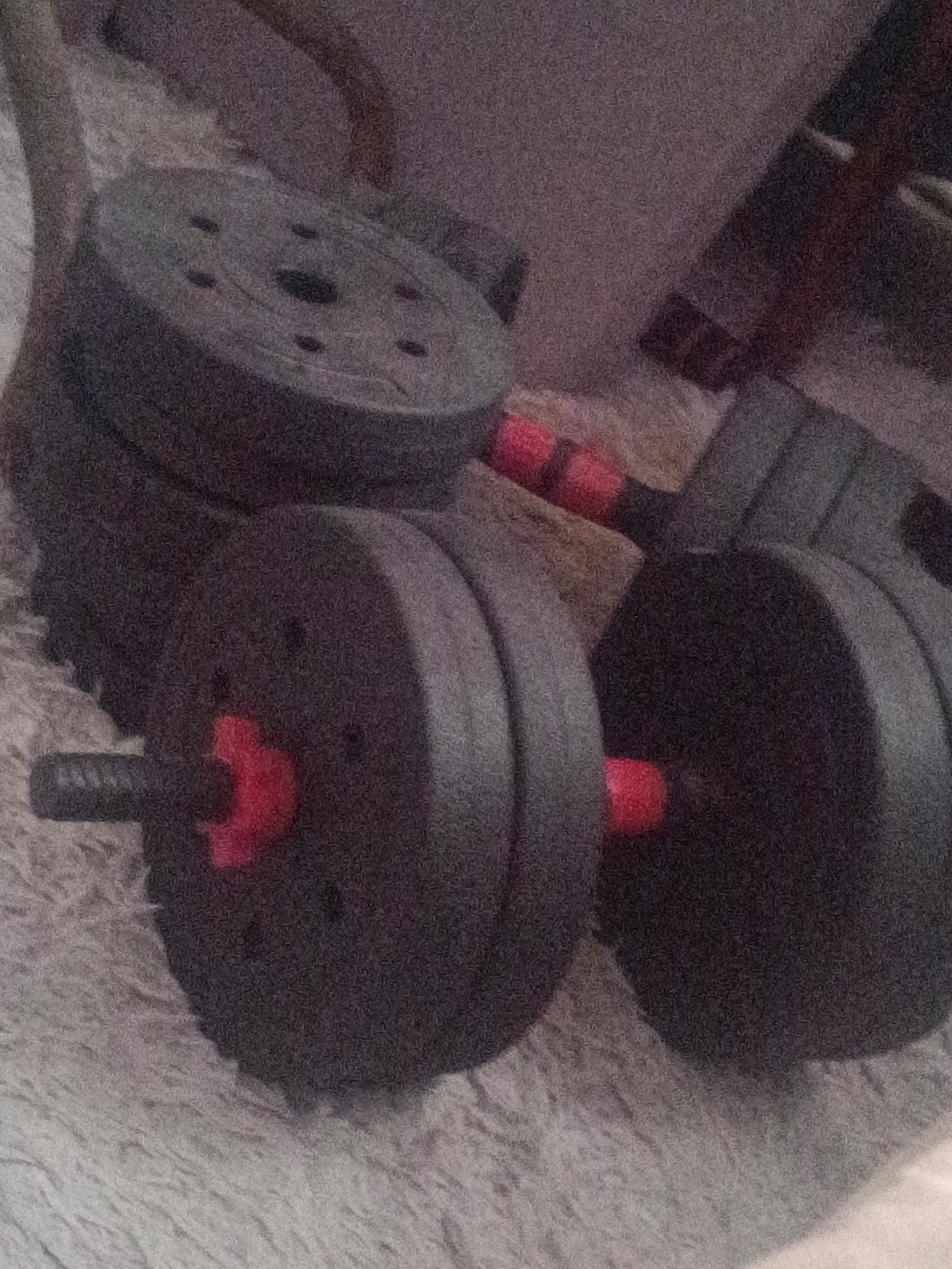 Two adjustable dumbbells are arranged on a textured, light-colored surface. Each dumbbell has a black handle with red accents at the ends, and multiple black weight plates of varying sizes. The weight plates appear to be made of metal, with a central hole for attachment to the handle. The scene is dimly lit, and the image quality is grainy. The background is blurred and indistinct, suggesting it is an indoor setting, possibly a home. No people are visible in the image. The location is Unknown, Unknown.