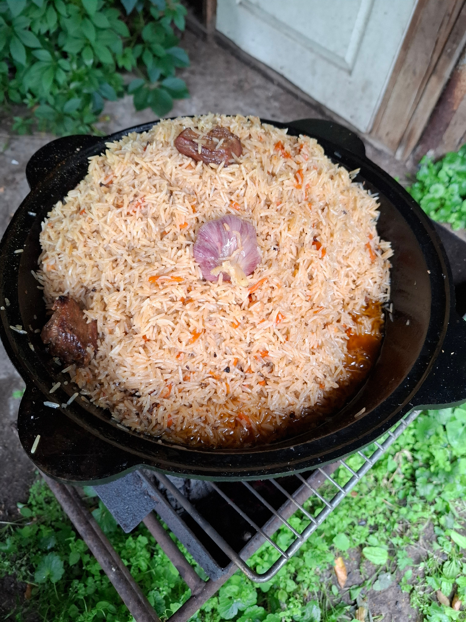 A close-up, overhead shot captures a large, black kazan (a traditional cast-iron cooking pot) filled with a heaping portion of pilaf. The pilaf is a light tan color, interspersed with small flecks of orange from carrots and darker specks, likely spices. Partially submerged in the rice are pieces of cooked meat and a whole head of garlic with its purple outer skin still on. The rice appears moist and slightly oily.
The kazan itself is set within a rustic metal grill or brazier, with embers visible beneath. The grill has a thick, dark metal construction with a crossbar design. The surrounding environment suggests an outdoor setting. Lush green foliage, typical of a garden or yard, occupies the background and spills into the foreground on the left side. The ground appears to be a mix of dirt and scattered leaves. A weathered wooden structure, possibly a fence or wall, is visible in the upper left corner.
The lighting is somewhat diffused, suggesting it could be daytime, possibly overcast or late afternoon, as there are no harsh shadows. The overall impression is one of rustic, outdoor cooking, perhaps a meal prepared in a traditional manner. There is no visible text in the image.