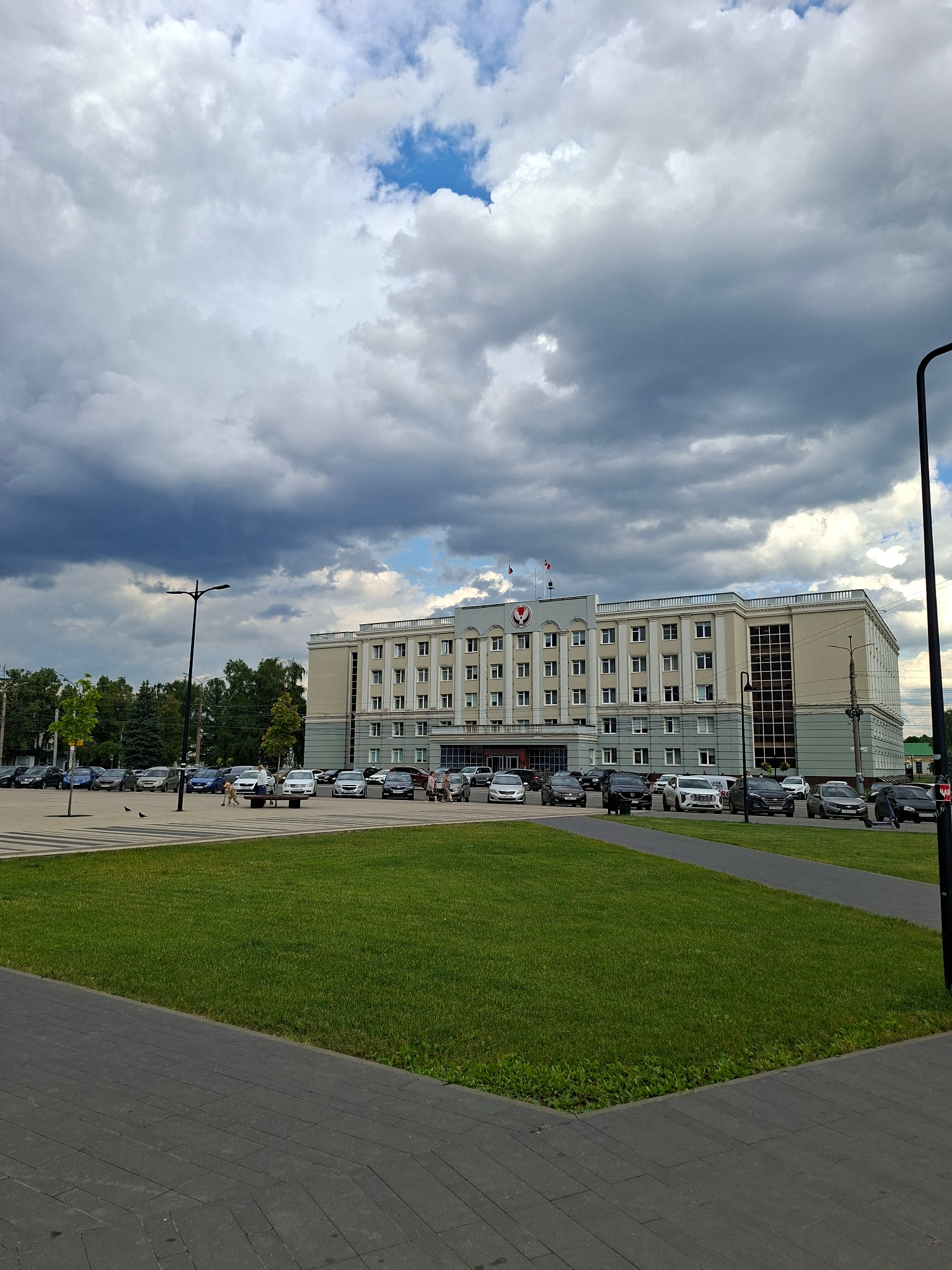 The image shows a modern building with a light-colored facade and multiple windows, situated next to a neatly manicured green lawn and a paved walkway. The building appears to be a public or administrative building, with flags flying near its entrance. A line of cars is parked along the side of the building, suggesting a place of work or public access.
The sky is filled with dramatic clouds, ranging from bright white to dark gray, indicating a dynamic weather system with potential for rain. Sunlight is breaking through in some areas, creating a play of light and shadow. A lamppost stands on the walkway, and a few pigeons are visible on the pavement.
In the distance, trees are visible, hinting at a semi-urban or suburban setting. A few people are walking on the walkway, some appearing to be interacting or walking dogs, suggesting a normal day. The overall scene conveys a sense of civic presence and everyday activity under a changeable sky. The time of day appears to be late afternoon or early evening, given the quality of the light and the cloud cover.