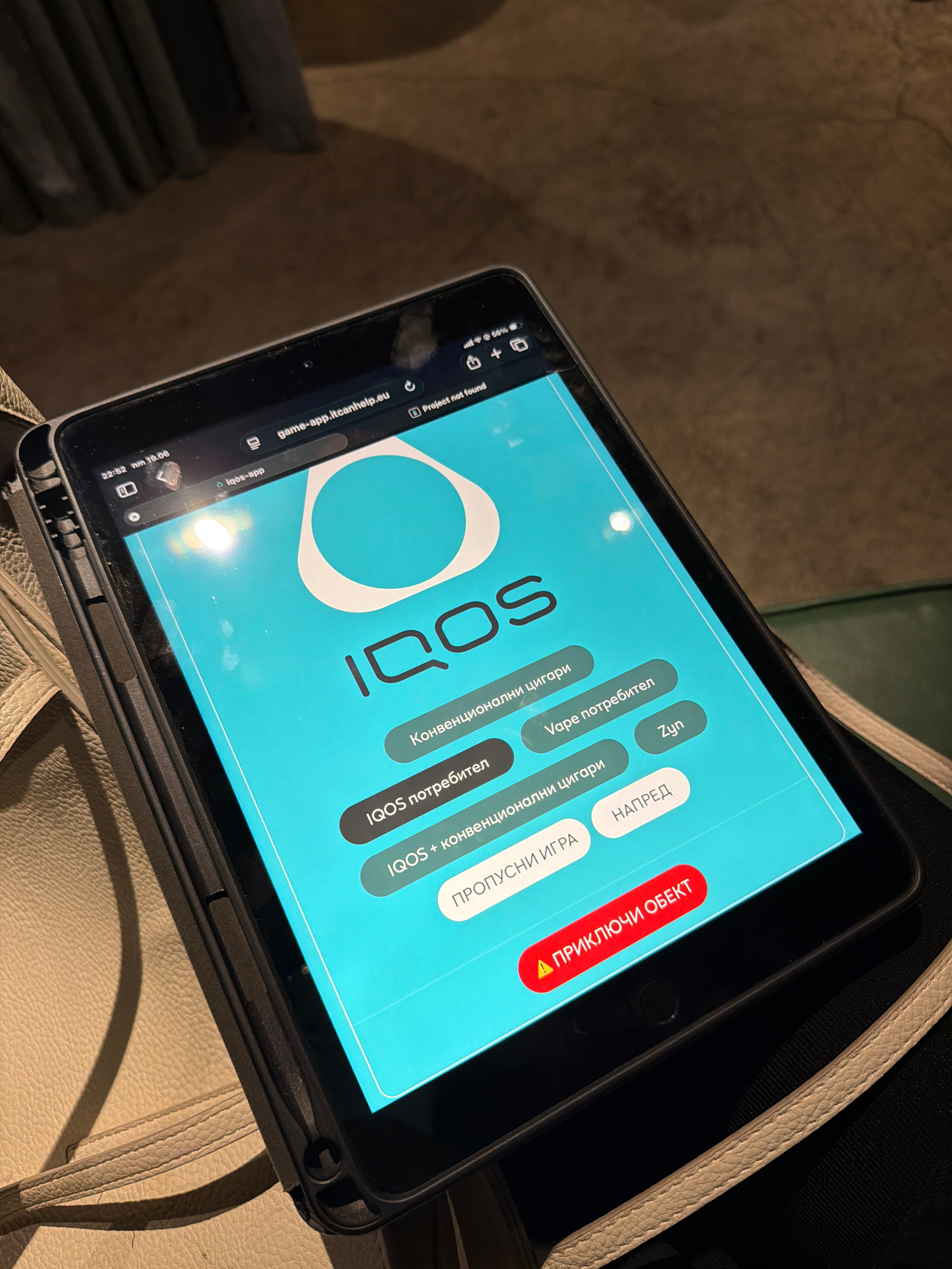 The image shows a tablet computer displaying a website with the URL "game-app.itcanhelp.eu". The screen has a turquoise background and features the IQOS logo, followed by several circular buttons with text in what appears to be Cyrillic script and English. Some of the visible text includes "IQOS", "Конвенционални цигари", "Vape потребител", "IQOS потребител", "Zyn", "IQOS + конвенционални цигари", "НАПРЕД", and "ПРИКЛЮЧИ ОБЕКТ". The tablet is placed on a surface that appears to be a bag or purse, made of light-colored textured material. The background is dark and out of focus, suggesting an indoor setting at night or in low light conditions. There are no people visible in the image. The time displayed on the tablet's status bar is "22:52". The location of Burgas, Bulgaria is not indicated in the image itself.