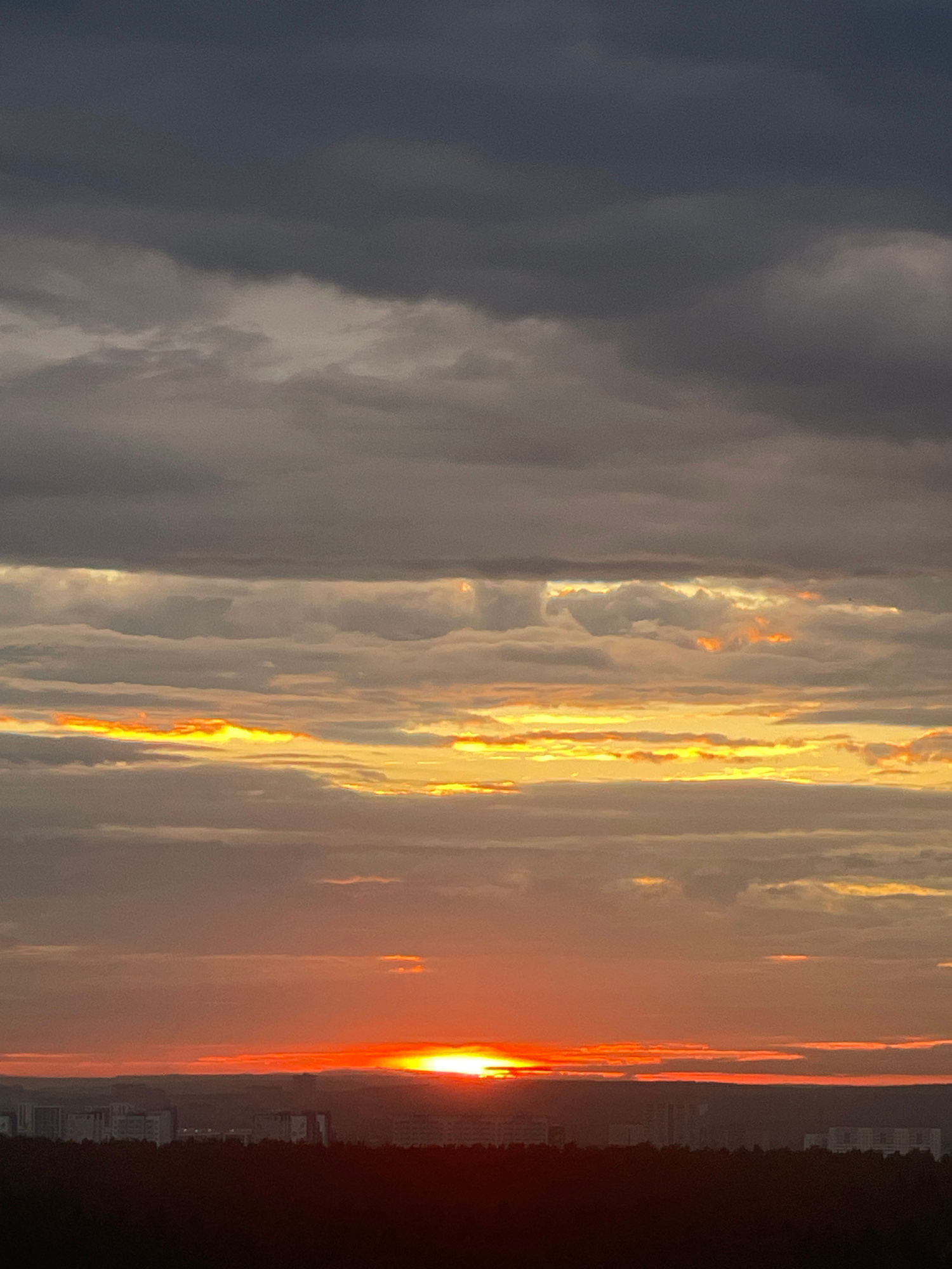 A vertical, high-angle shot captures a dramatic sunset over a city, likely Perm, Russia. The sky is layered with clouds, transitioning from dark gray at the top to lighter gray and orange hues closer to the horizon. The sun, a brilliant orange orb, is partially obscured by clouds just above a distant, dark silhouetted treeline and city skyline. The city buildings are small and indistinct against the darkening landscape. The overall impression is one of a moody and atmospheric twilight.