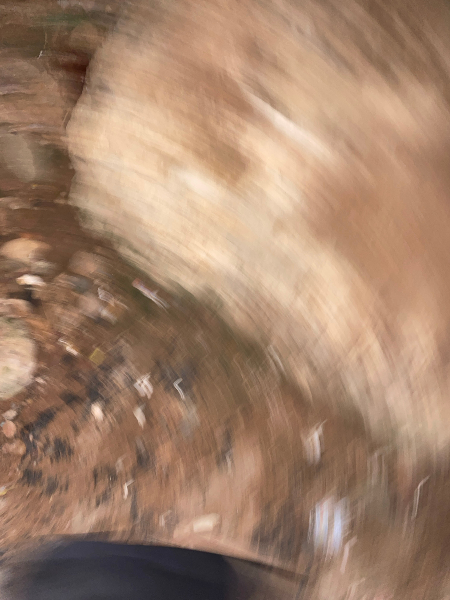 This is a blurry, abstract image taken from a high angle, likely outdoors in Lokoja, Nigeria. The foreground is dominated by a dark, out-of-focus shape, possibly clothing or a surface. The background depicts a textured, earthy ground with scattered debris, including small rocks and what appear to be pieces of white and colored trash. A large, light-colored, indistinct form with blurred vertical striations occupies the upper right portion of the frame, suggesting a rock formation or a large object. The overall impression is of movement and chaos due to the motion blur.