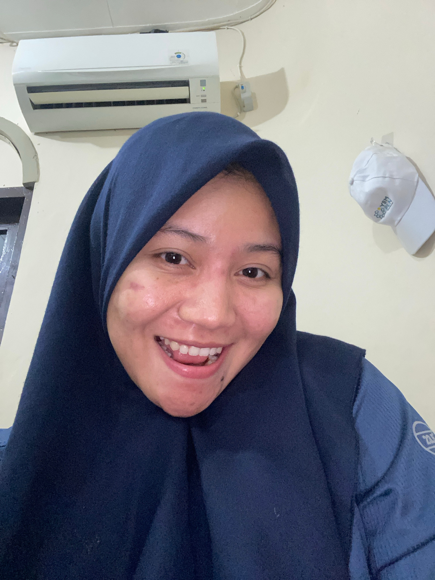The image features a close-up, portrait-style selfie of a young woman wearing a dark blue hijab and a matching dark blue garment, likely a jacket or sweater. She is smiling broadly, with her mouth open, revealing her teeth. Her eyes are open and looking directly at the camera. The background is a light-colored wall, with an air conditioning unit mounted on it to the left of the frame. Above and to the left of the woman's head, a white baseball cap with some text and a logo is visible, hanging on the wall. The lighting suggests an indoor setting, possibly during the daytime. The woman appears happy and relaxed. The visible text on the cap is "ROBIN" and "YBM". The overall impression is that of a casual, personal photograph taken in a home or indoor environment in Banjarbaru, Indonesia.