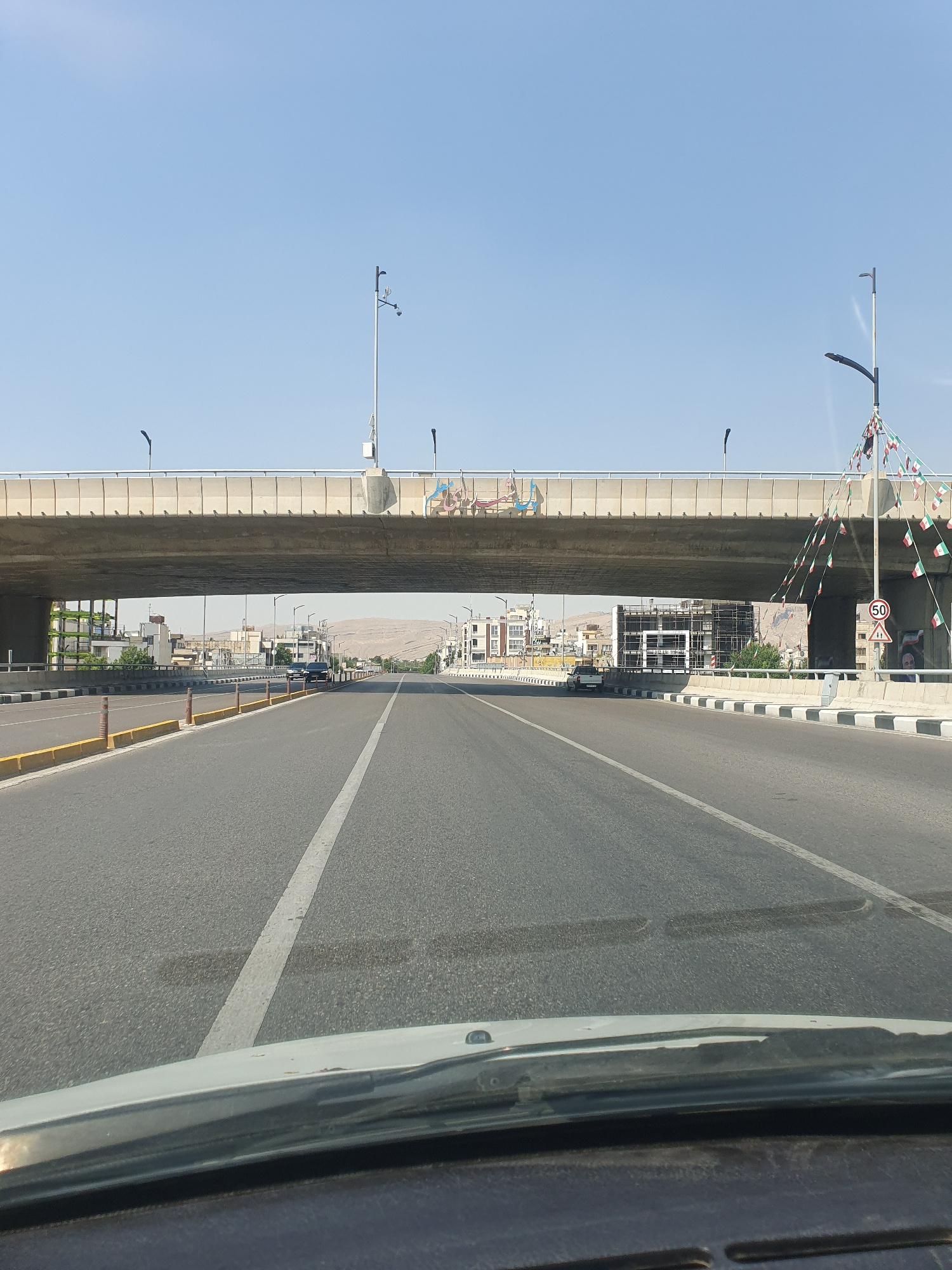 The image shows a view from inside a vehicle, looking forward on a multi-lane road. A large concrete overpass dominates the upper portion of the frame, casting a shadow over the road below. The overpass is decorated with numerous small flags, appearing to be Iranian flags. On the right side of the overpass, a lamppost is visible with flags draped around it and a speed limit sign indicating "50".
The road itself is asphalt with clear lane markings. There are barriers along the sides of the road, some with yellow markings. On the left side of the road, there are buildings and trees visible, suggesting an urban or suburban environment. On the right side, beyond the barriers, more buildings and a distant, arid mountain range can be seen. A few cars are present on the road, including a dark SUV under the overpass and a white car further down the road. The sky is clear and blue, indicating a daytime scene.
While the image does not explicitly show landmarks that pinpoint it to a specific location within Shiraz, Iran, the style of the overpass, the flags, the architecture of the buildings, and the presence of the speed limit sign (likely in Persian script, although the script is not clearly legible) are consistent with urban infrastructure found in Iran. The mountainous backdrop is also characteristic of the landscape surrounding Shiraz. Without further contextual information or clearer identifiable landmarks, a precise location within Shiraz cannot be determined.