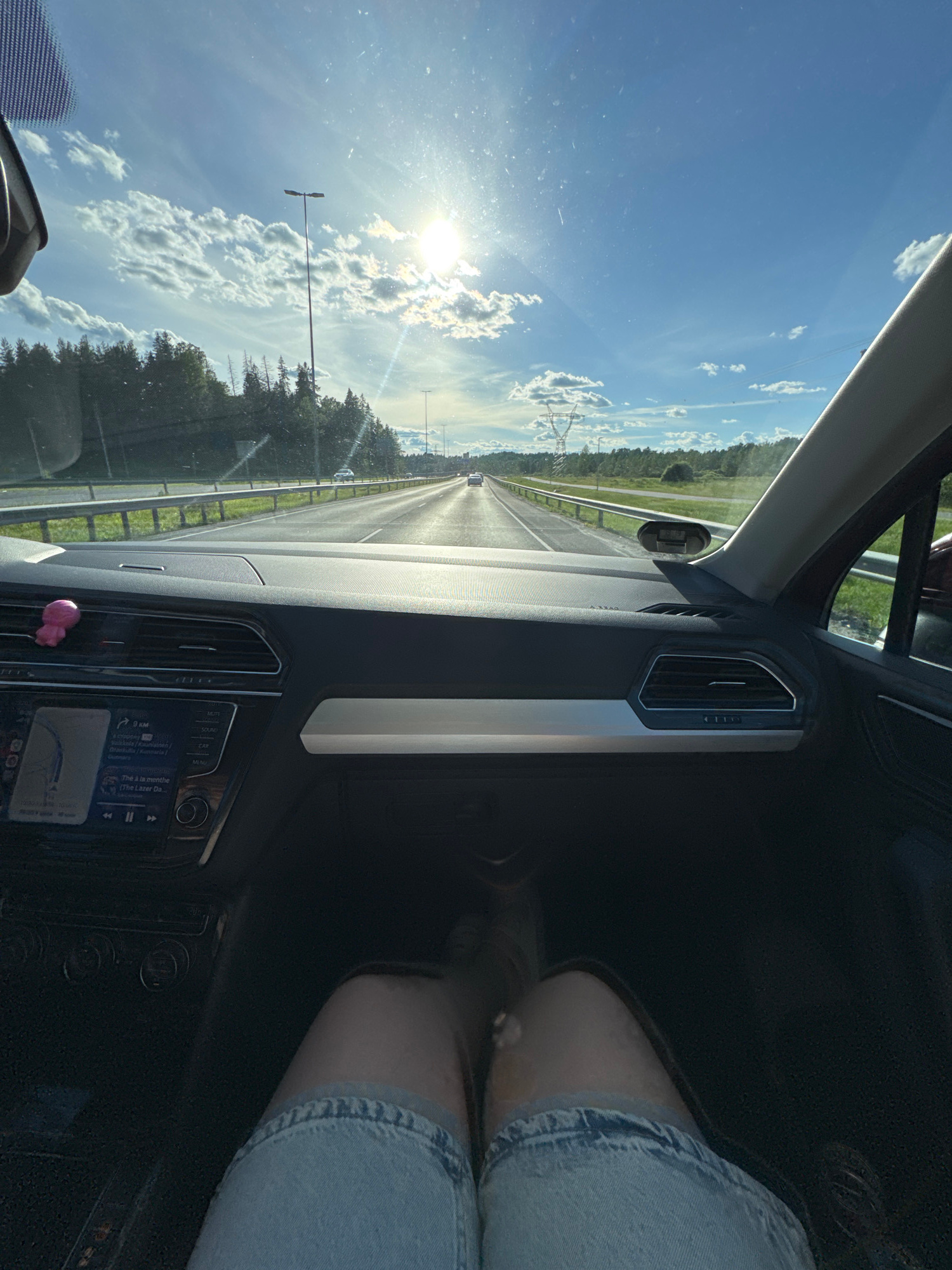 The image captures a view from inside a vehicle, looking forward through the windshield. The sun is high in a bright blue sky with scattered clouds, casting a glare on the windshield. The road ahead is a highway with multiple lanes, bordered by a metal guardrail and a grassy shoulder. Trees line the sides of the road, and tall lamp posts are visible along the highway. The dashboard of the car is in the foreground, with a GPS navigation system displaying a map. A small pink object is attached to the dashboard. The lower portion of the image shows a person's legs, wearing denim shorts. The scene appears to be a daytime drive on a highway.