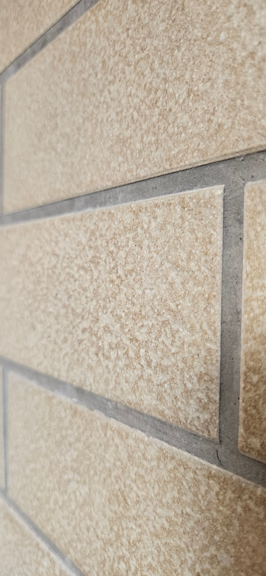 The image shows a close-up, angled view of a wall tiled with rectangular, light brown tiles that have a speckled texture. The tiles are arranged horizontally, with thin, dark gray grout lines separating them. The perspective is shallow, with some tiles in focus and others blurred due to the close proximity and angle. The lighting appears to be natural, casting subtle shadows and highlighting the textured surface of the tiles. There are no people or other discernible objects in the frame.
