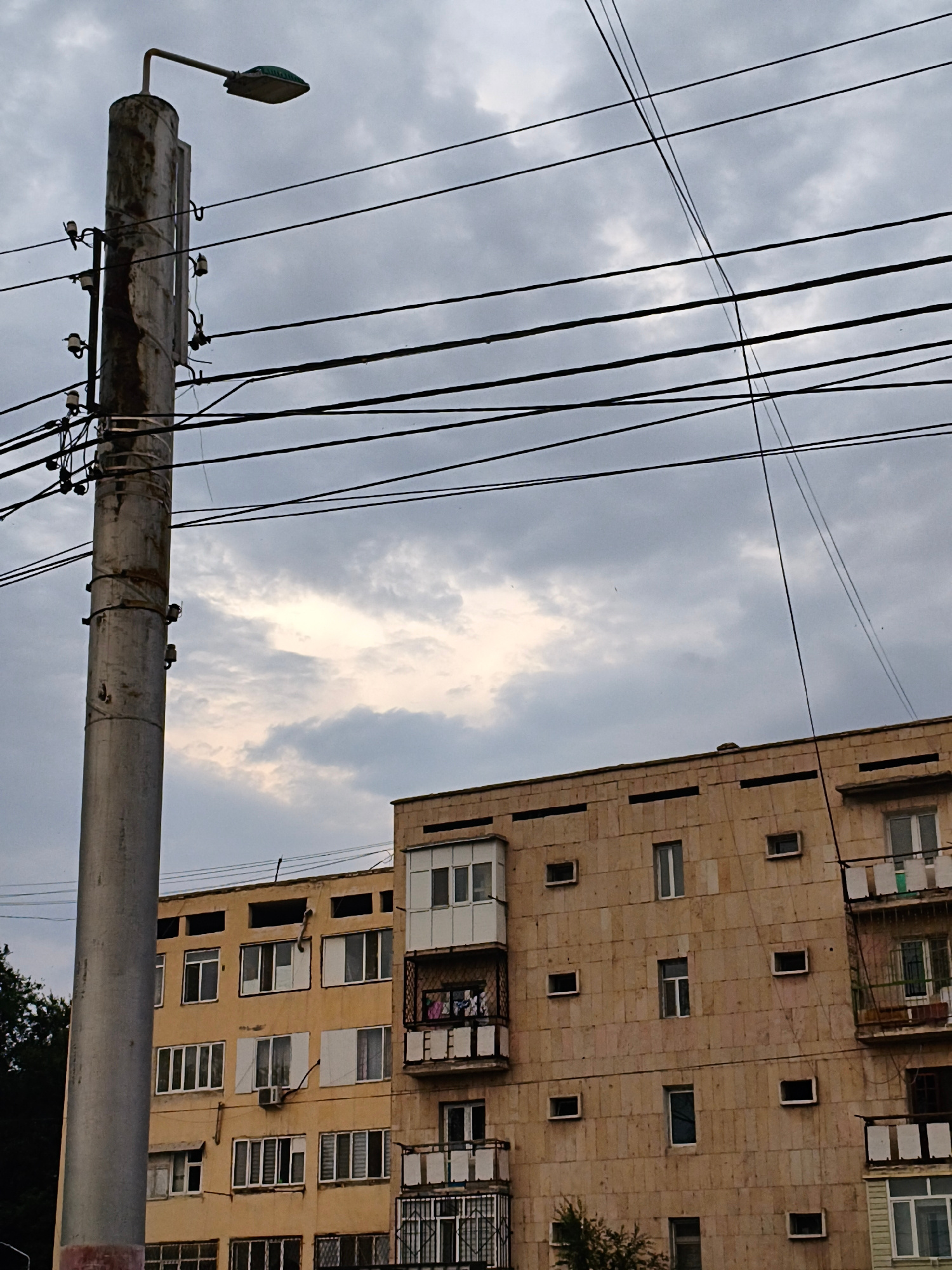 This media file captures a slightly overcast sky with dramatic clouds, suggesting a late afternoon or early evening setting. Dominating the left side of the frame is a weathered concrete utility pole, adorned with electrical insulators and a street lamp at its apex. A dense network of dark power lines crisscrosses the sky above a pale yellow apartment building. The building itself exhibits a uniform, somewhat drab facade with numerous windows of varying sizes. Some windows are covered with white blinds, while others reveal glimpses of indoor spaces. Balconies with metal railings are visible on the right side of the building, with laundry hanging to dry on one of them, hinting at the presence of inhabitants. The architectural style and color of the building, along with the overall atmosphere, evoke a sense of a residential area in a post-Soviet urban landscape. There are no visible people, text, or specific landmarks to pinpoint an exact location beyond the general urban setting. The image conveys a sense of everyday life within a city, under a moody sky.