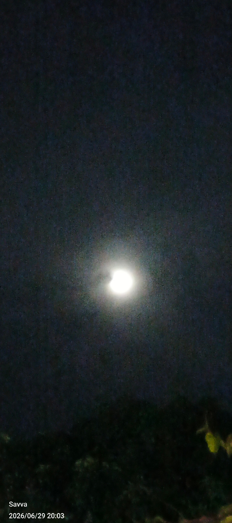 The image depicts a partial solar eclipse at night, with the moon obscuring a portion of the sun. The sun is a bright, circular object with a dark crescent shape on its left side, indicating the eclipse. The sky is dark and appears to be nighttime. In the foreground, there is a silhouette of trees and foliage, suggesting an outdoor setting. The lower left corner of the image contains visible text: "Savva" and "2026/06/29 20:03". The date and time indicate the image was captured on June 29, 2026, at 8:03 PM. The location context is Phu Thuy Ward, Vietnam. The scene is dark and serene, likely capturing a celestial event.