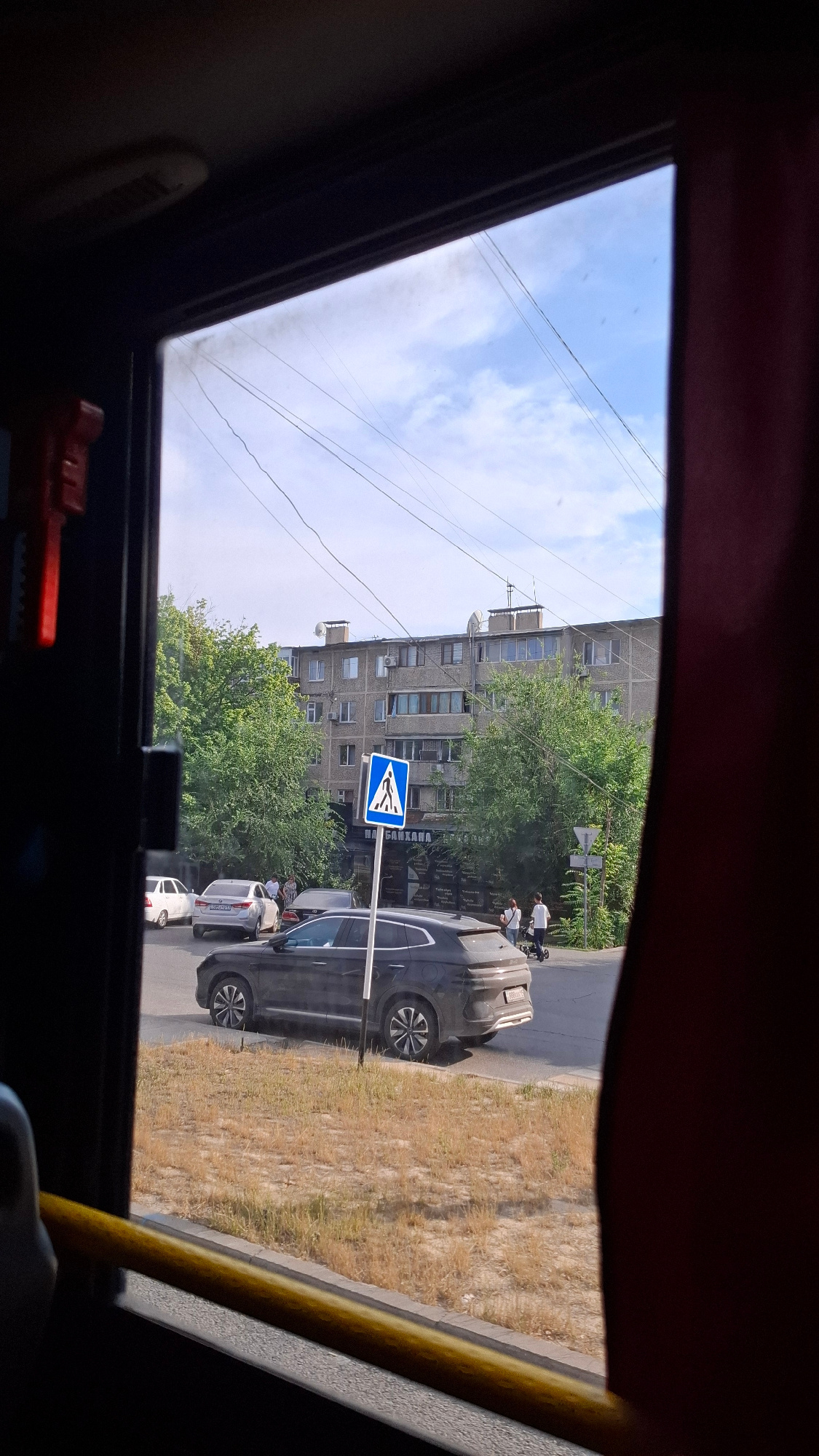 The image captures a view from inside a vehicle, likely a bus, looking out through a sunroof or large window. The scene is oriented vertically, suggesting the photo was taken while the vehicle was moving or held at an angle.
The primary subjects are urban elements: a multi-story apartment building, a street with parked cars, and pedestrians. The building appears to be made of concrete panels, typical of Soviet-era architecture. In front of the building, a pedestrian crossing sign, a blue square with a white pictogram of two people walking, is prominently displayed. Several cars are visible, including a dark-colored SUV-like vehicle and a lighter-colored sedan.
Two individuals, dressed in white, are seen on the sidewalk near the pedestrian crossing, one pushing a stroller or cart. The dry, overgrown grass along the side of the road suggests a warm climate. Power lines crisscross the sky, which is partly cloudy with a bright blue expanse. The lighting indicates it is daytime, likely mid-morning or afternoon given the bright sunlight.
A yellow pole, possibly part of the vehicle's interior, runs along the right edge of the frame, reinforcing the perspective from within. The overall atmosphere is one of everyday urban life. There is some visible text on a sign on the building, but it is not clearly legible. The setting appears to be Shymkent, Kazakhstan, as indicated by the location context.