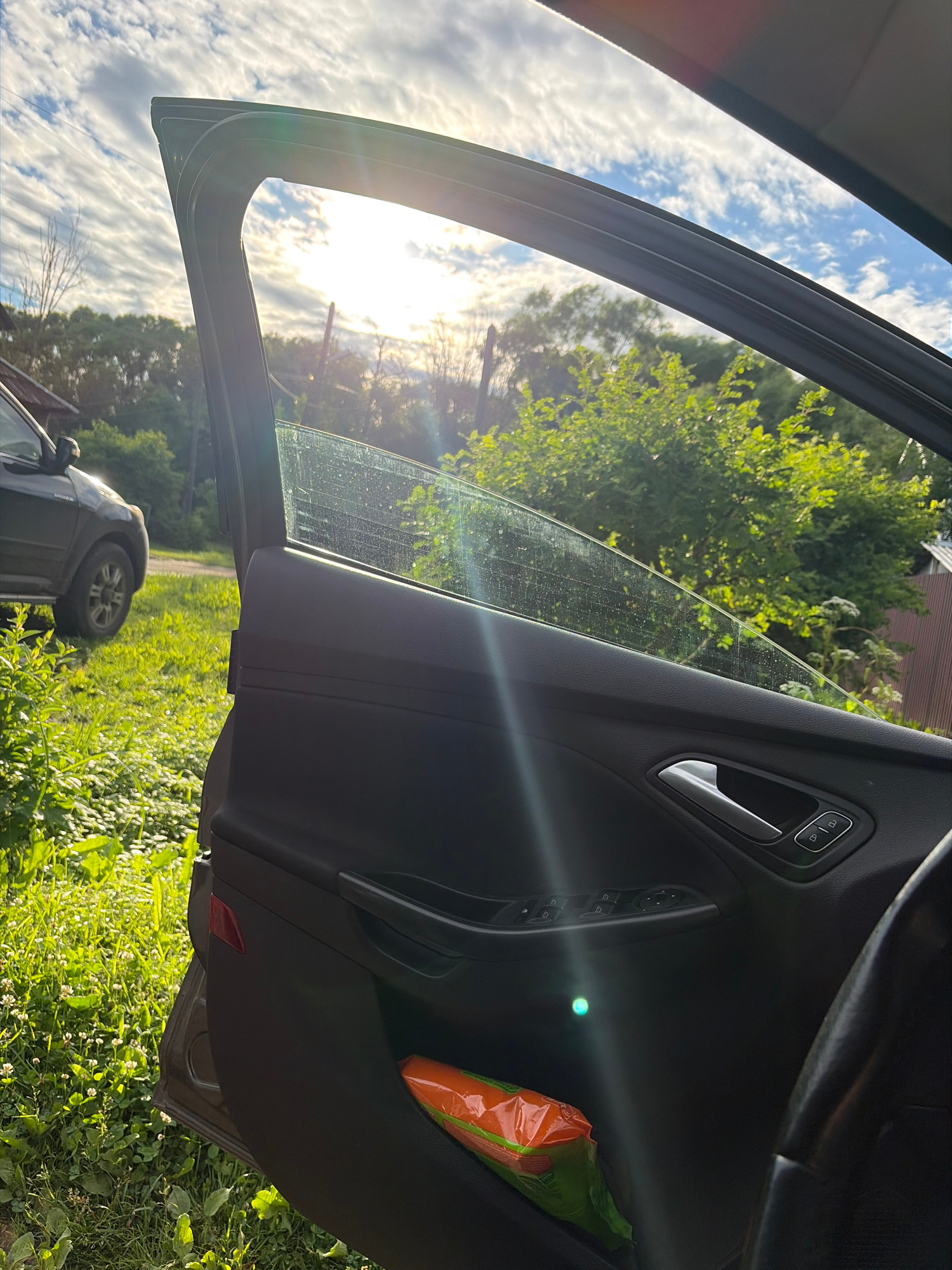 The image captures a view from inside a car, looking out through the open driver's side door. The door panel is visible in the foreground, featuring a silver door handle and buttons for window controls and door locks. A bright orange package, possibly containing food or supplies, is tucked into a compartment on the door. Sunlight streams in from the left, creating lens flare and illuminating the car's interior.
Through the open door and window, a sun-drenched outdoor scene unfolds. The sky is a vibrant blue with scattered white clouds. Lush green trees and foliage dominate the background, suggesting a natural or semi-rural setting. The sun appears to be positioned low in the sky, casting long shadows and indicating either early morning or late afternoon. The overall impression is one of a bright, sunny day, likely in a temperate climate.
In the lower portion of the image, the rear wheel and part of the exterior of another vehicle are visible. This suggests the car the photo is taken from is parked, possibly on a grassy or unpaved area, as indicated by the green vegetation and the presence of small white flowers on the ground. The light quality and the surrounding greenery suggest a warm season, likely summer. No people are visible in the image. The image appears to be taken in Kimry, Russia.