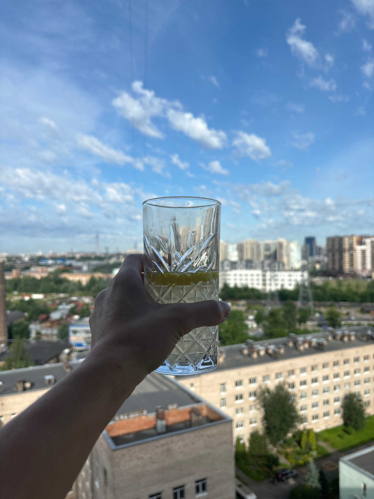 A hand holds a glass of beverage with a lemon slice against a blue sky with scattered clouds. In the background, a cityscape unfolds with buildings of various heights, including apartment complexes and some industrial structures. Trees and greenery are visible between the buildings, suggesting a suburban or partially developed urban area. The perspective is from a high vantage point, likely an upper floor of a building, overlooking the surrounding city.