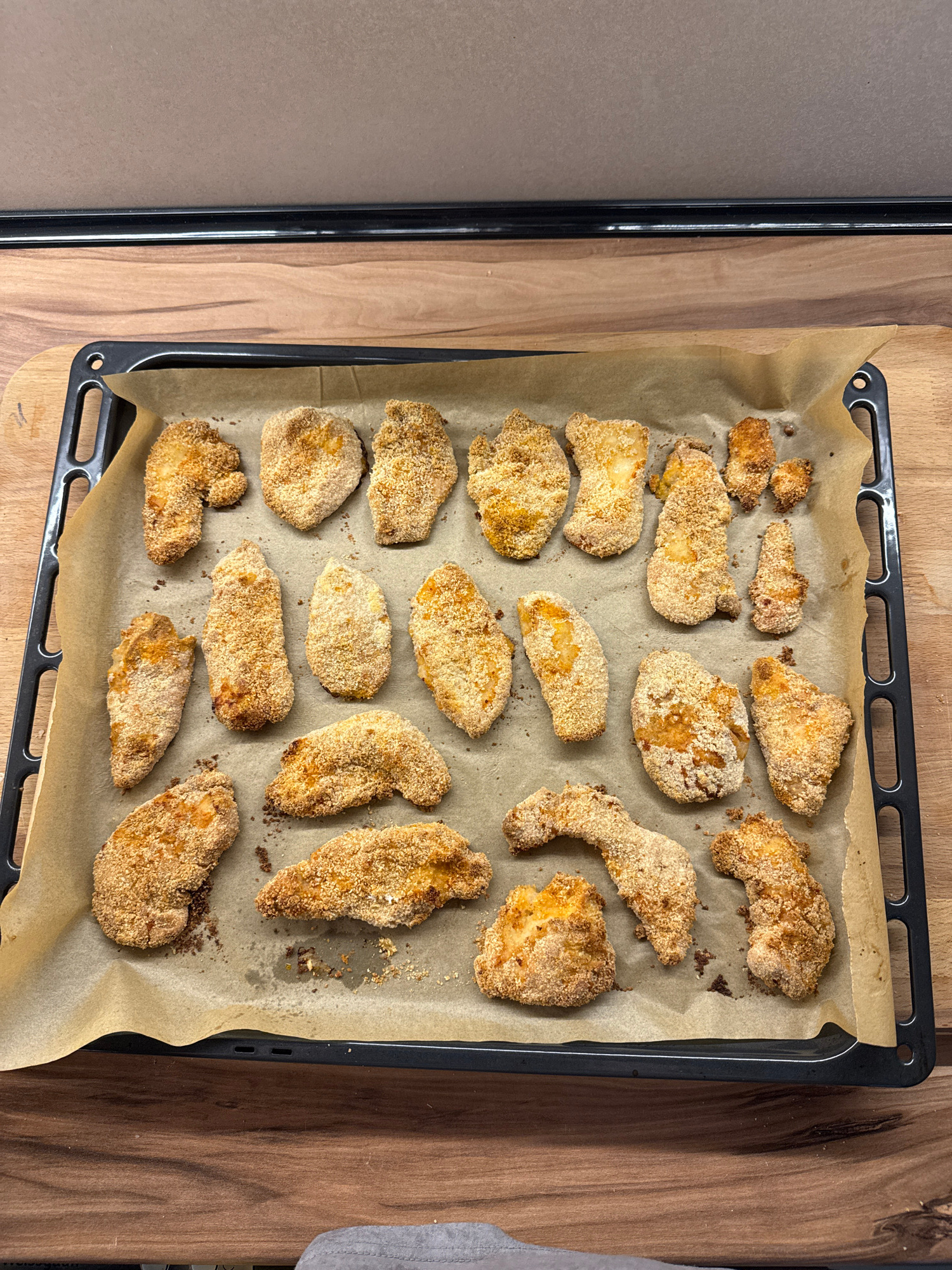 The image shows a baking sheet lined with parchment paper, filled with breaded chicken pieces. The chicken pieces are golden brown and appear to be cooked. They are arranged in a somewhat scattered pattern on the baking sheet. The baking sheet itself is black and has a slightly raised edge. It rests on a wooden surface, possibly a countertop or a table. The background shows more of the wooden surface, with a vertical black object on the left side of the image, which could be part of a kitchen appliance or cabinet. The lighting suggests an indoor setting, likely a kitchen, with diffused light that doesn't cast harsh shadows. There are no people visible in the image. The overall impression is of food preparation or serving, with the chicken pieces ready to be eaten or further processed. The breading on the chicken looks crispy, suggesting they might have been baked or fried. The image is taken from a top-down perspective, providing a clear view of the entire baking sheet and its contents.