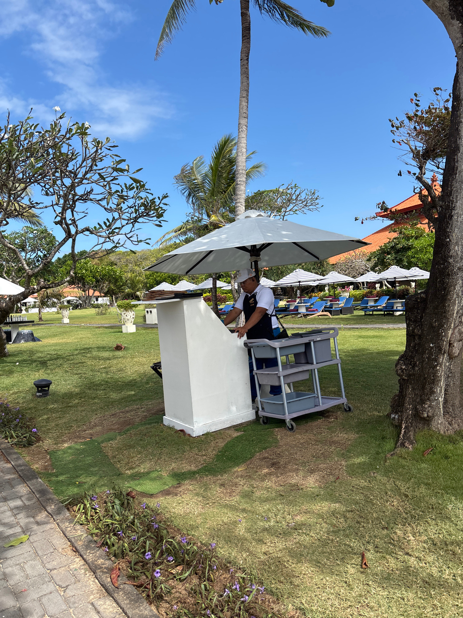 A man wearing a white cap and shirt with a dark apron stands at a white counter outdoors in Kuta Selatan, Indonesia. He is facing a grey, wheeled cart with three shelves, which is positioned next to the counter. The counter is partially covered by a large white umbrella. In the background, there is a lush green lawn with several lounge chairs and umbrellas set up. Palm trees and other tropical foliage are visible, along with a building with an orange tiled roof. The sky is clear and blue with some scattered clouds. The man appears to be attending to the cart or counter.