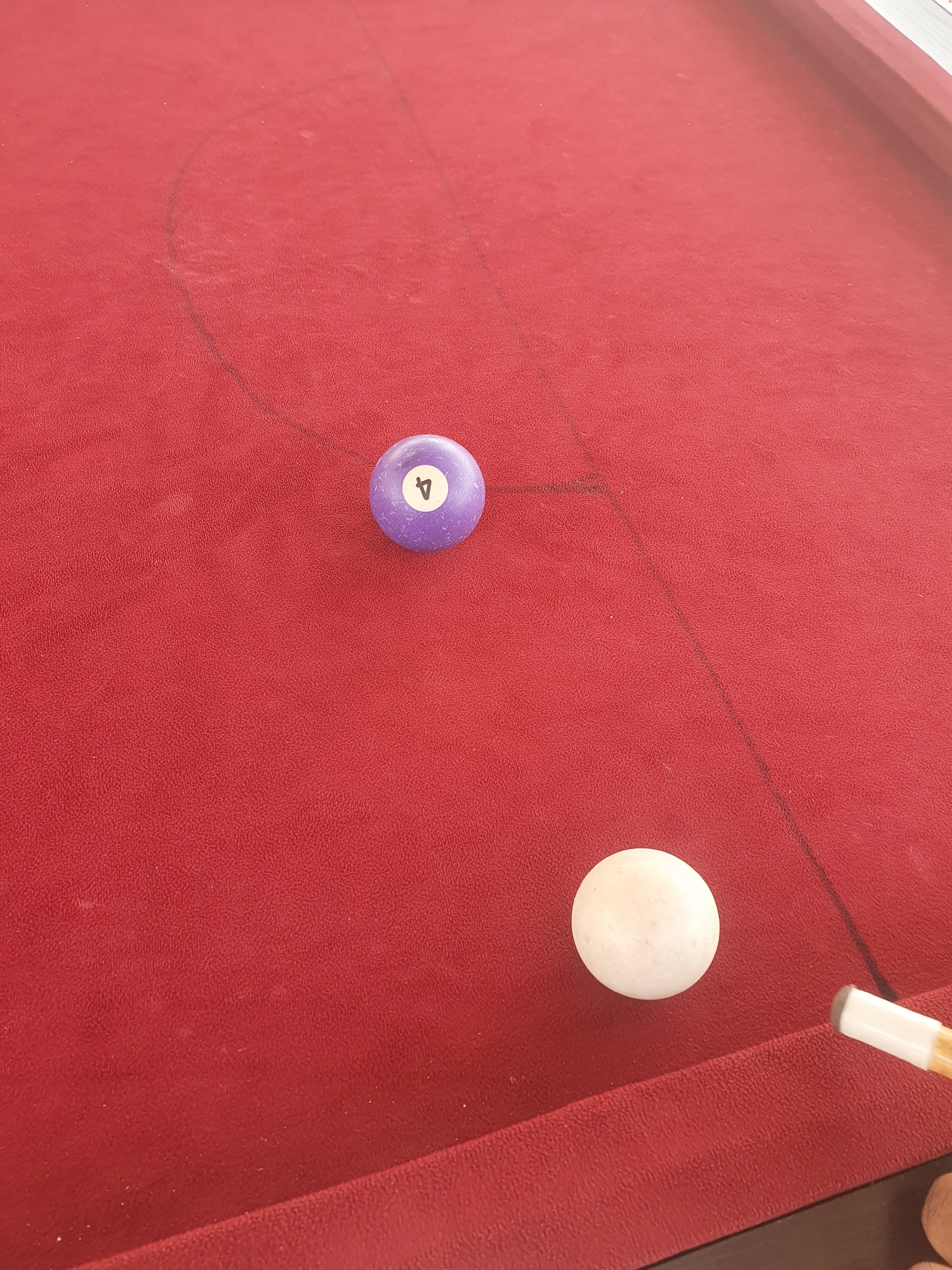 The image depicts a close-up, high-angle view of a red billiards table. Two pool balls are visible: a purple ball with the number "4" on it, and a white ball. A portion of a pool cue stick is also visible in the lower right corner, positioned near the white ball, suggesting an imminent shot. Faint chalk marks on the felt indicate the playing area, with a curved line and a straight line visible. The lighting suggests an indoor environment, possibly outdoors on a covered patio due to the bright, natural-looking light. There are no people or animals visible in the frame. The scene is static, capturing a moment before a billiard shot. The location of Lokoja, Nigeria is not depicted in this image.