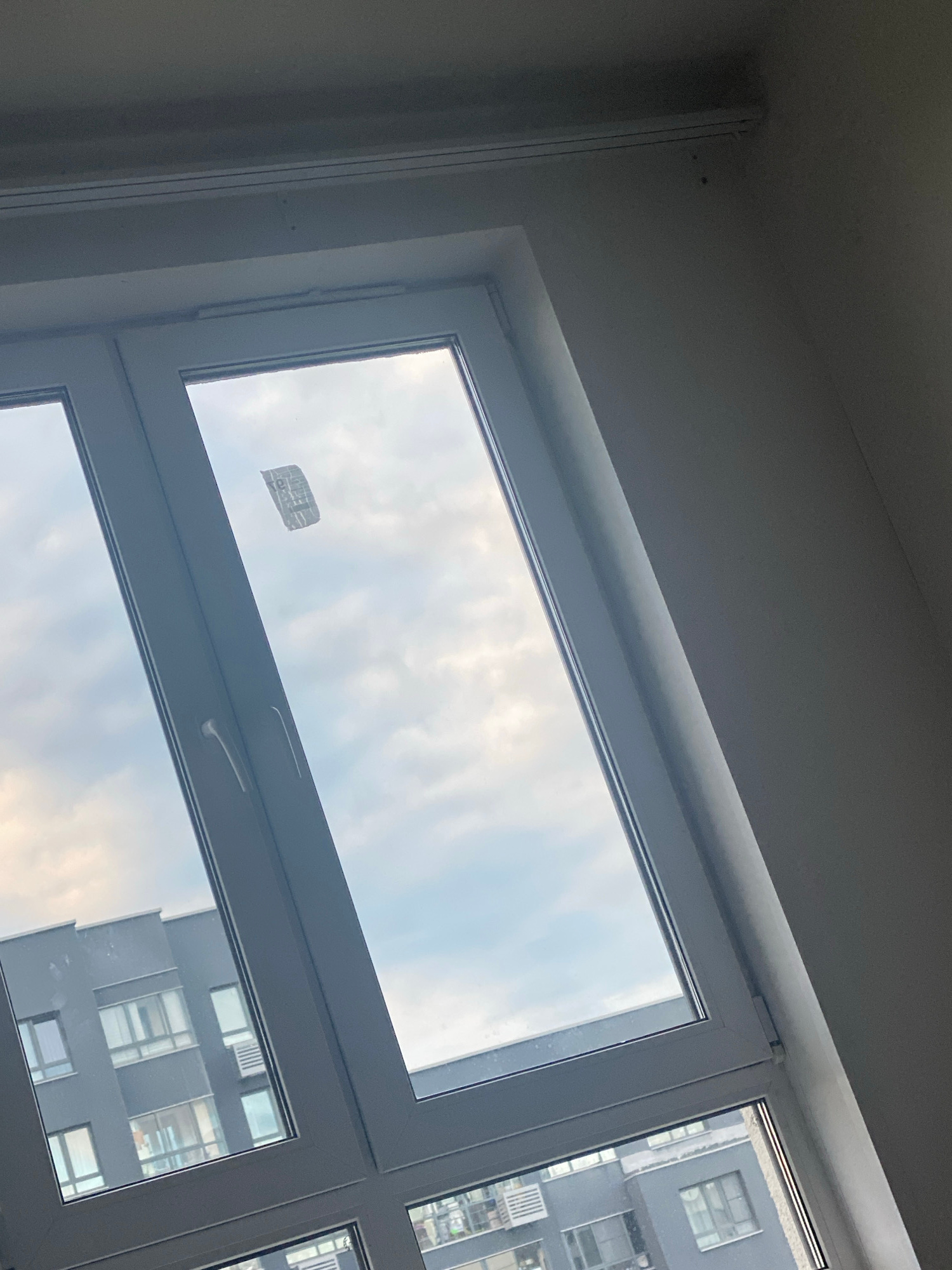 A close-up, angled shot from inside a room shows a view through a white-framed window. The window is divided into two panes by a vertical mullion. The left pane is partially visible and shows a modern, grey apartment building with multiple windows and balconies. The right pane, which is more prominent, looks out onto a cloudy sky. A small, crumpled piece of what appears to be paper or plastic is stuck to the inside of the right window pane, near the top. The wall of the room is visible on the right side of the image, appearing to be painted a light, neutral color. The angle of the shot suggests the camera is positioned relatively low and looking upwards.