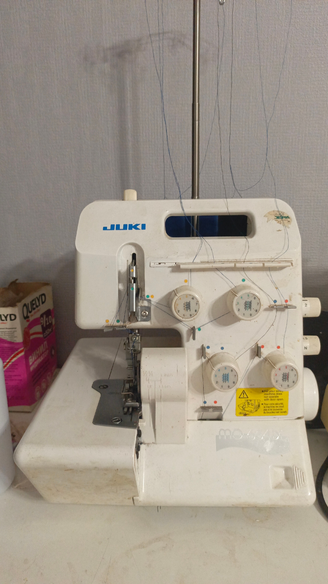 A white JUKI serger sewing machine is positioned on a white surface, likely a table, against a textured gray wall. The machine is threaded with blue thread, which hangs loosely from spools above. The serger has several dials and levers for adjusting stitch settings. To the left of the machine, a pink and white box labeled "QUELYD" is partially visible. The environment appears to be an indoor space, possibly a room in Sertolovo, Russia, with some ambient lighting casting a shadow of the serger's thread guide onto the wall.