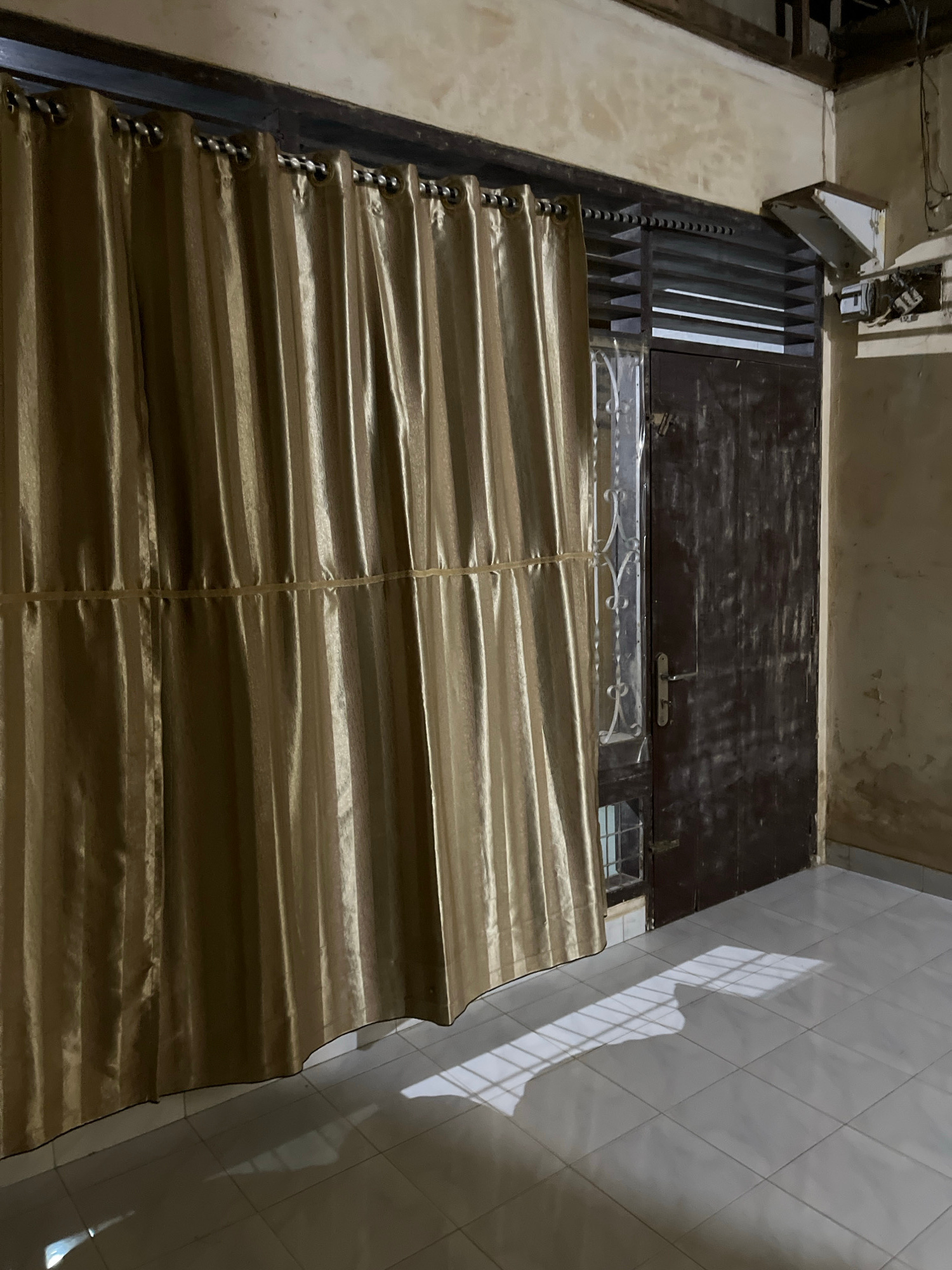 The image shows a view from inside a building looking out at a partially covered doorway or window. The foreground is dominated by a golden-colored fabric blind, which is pleated horizontally and appears to be made of a slightly shiny material. The blind is partially drawn, obscuring the view beyond. Above the blind, there is a dark-colored door or gate with vertical slats. To the left of the blind and gate, there is a textured, light-colored wall.
In the upper left portion of the image, there is an electrical junction box mounted on the wall, with wires visible and some damage or disrepair. The top of the gate appears to have some decorative metalwork.
To the right of the gate, a tiled wall is visible, with diamond-shaped tiles. Sunlight is casting a strong shadow of what appears to be a window with grilles onto this tiled wall, suggesting it is daytime.
The overall impression is an interior view of a room or hallway in a building, possibly a residential or commercial space, with a focus on the window covering and the adjacent architectural elements. There is no visible text in the image. The setting appears to be Kota Kuala Simpang, Indonesia, as per the context.