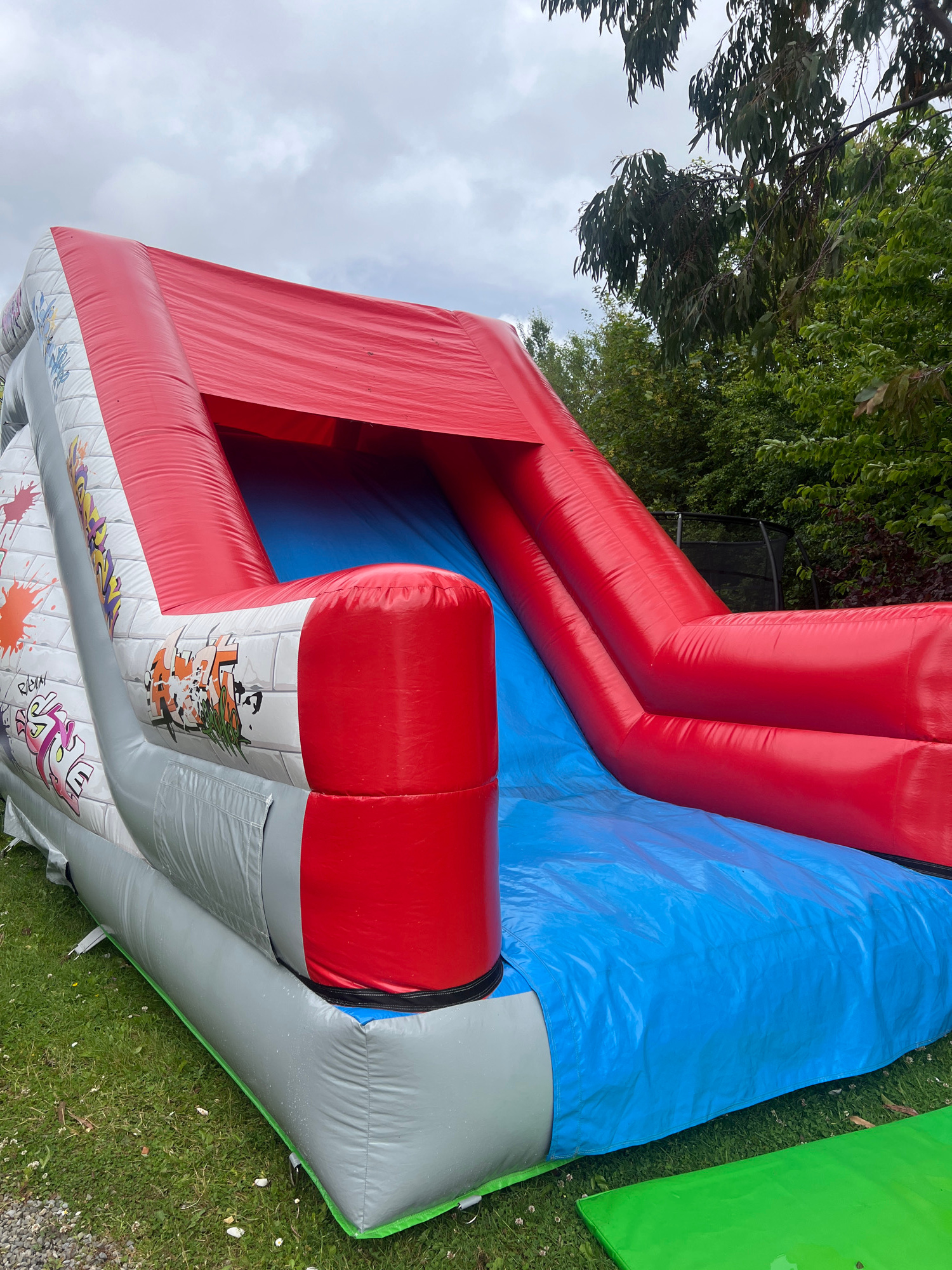 This media file shows a large, inflatable play structure in an outdoor setting. The structure is primarily red and blue, with some grey sections that feature colorful graffiti-style artwork. The artwork includes stylized text and cartoonish characters. The inflatable appears to be situated on a grassy area, with trees and foliage visible in the background. A portion of a green inflatable or barrier is also visible to the right of the main structure. The ground is covered in green grass with some small white flowers. The sky is overcast, suggesting a cloudy or possibly rainy day. The image is taken from a high angle, looking down and across the inflatable. There is no clear indication of people or a specific activity taking place, other than the presence of the play structure itself, which is typically used for recreation. The overall impression is of an outdoor event or setup for children's entertainment. The location context provided is Kilpedder, Ireland, which implies the scene is set in that region.