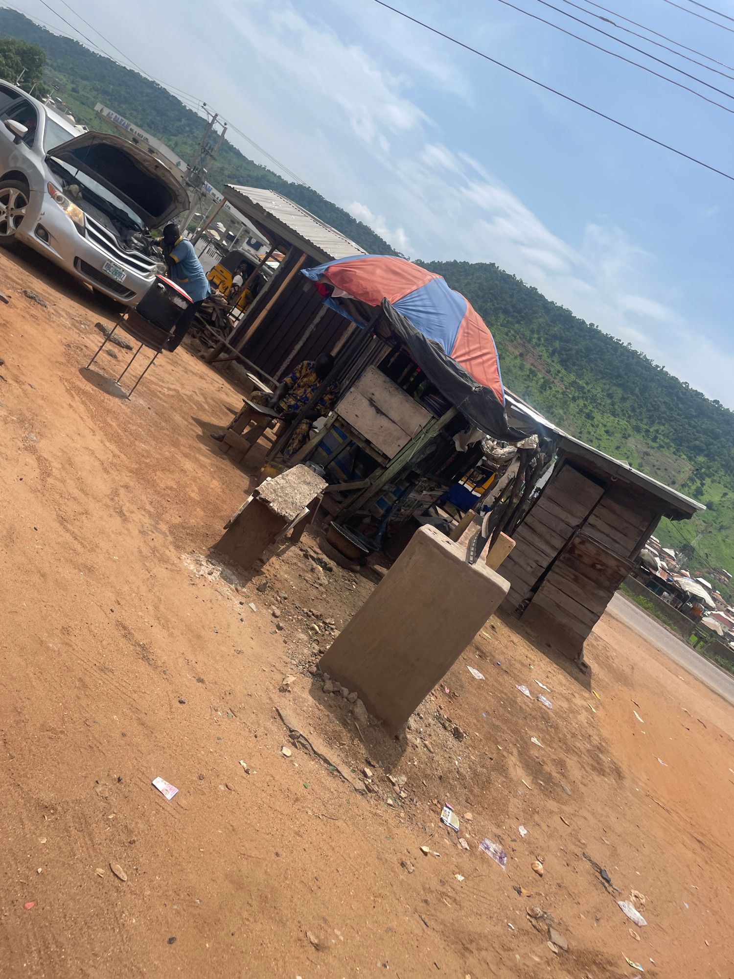 A silver car is parked with its hood open, suggesting a mechanical issue, while a person in a blue shirt works on it. Nearby, a roadside stall with a red and blue umbrella is set up. The stall appears to be made of wood and metal, with various items displayed. The ground is covered in reddish-brown soil, with scattered debris and a few pieces of paper money. In the background, a lush green hillside rises, with a town or village visible on its slopes. The sky is a clear blue with a few wispy clouds. This scene is consistent with a location in Lokoja, Nigeria, which is known for its hilly terrain and roadside commerce.