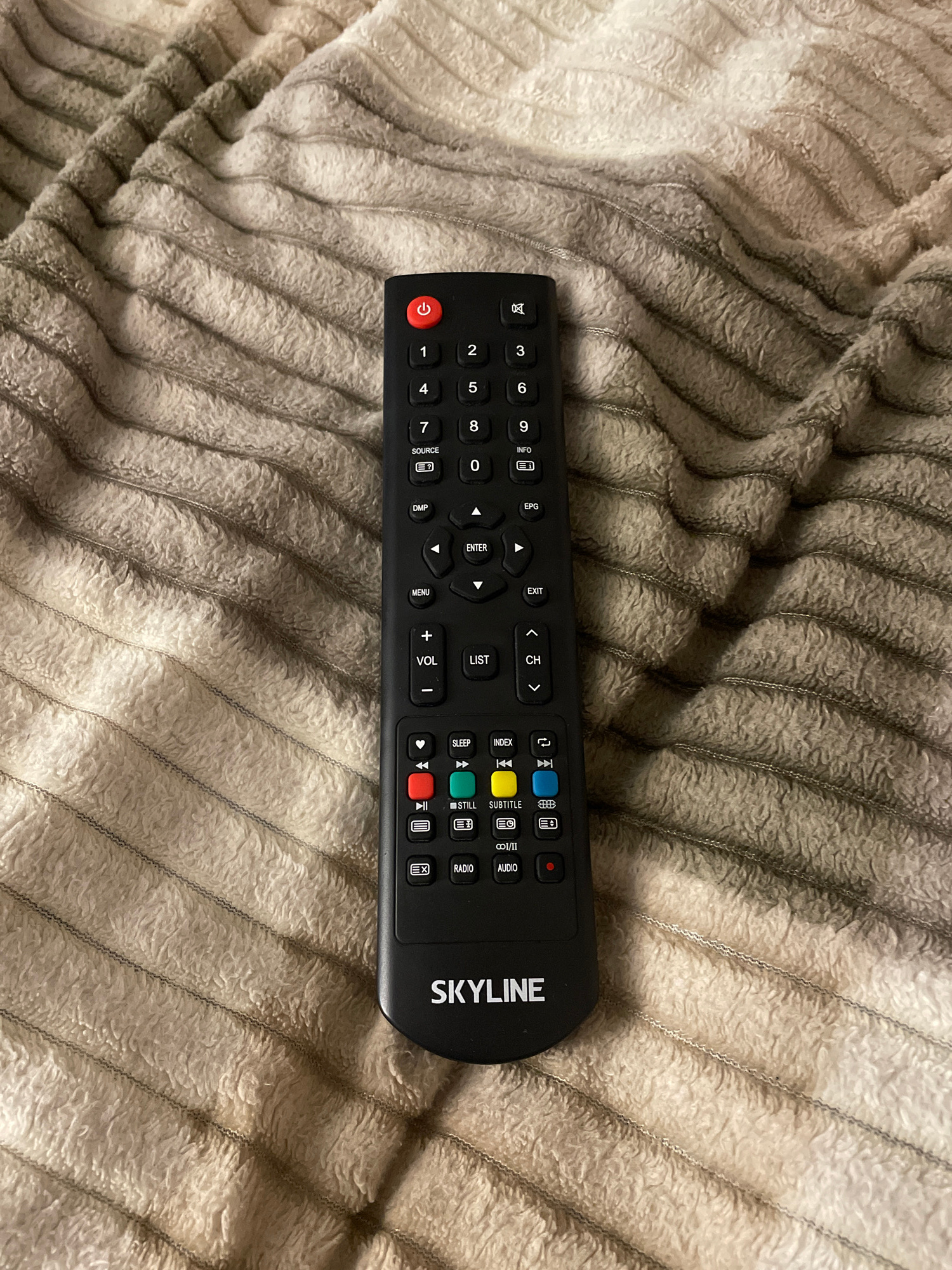 A black remote control rests on a textured, striped blanket. The remote has a red power button at the top, followed by a numerical keypad from 0 to 9. Below the numbers are various function buttons, including "SOURCE", "DMP", "ENTER", "MENU", "EXIT", "VOL", "LIST", and "CH". Further down are playback control buttons, color-coded buttons (red, green, yellow, blue), and buttons labeled "SLEEP", "STILL", "SUBTITLE", "CD1/II", and "AUDIO". The brand name "SKYLINE" is visible at the bottom of the remote. The blanket is a light beige color with darker beige stripes. The image is taken from a top-down perspective.