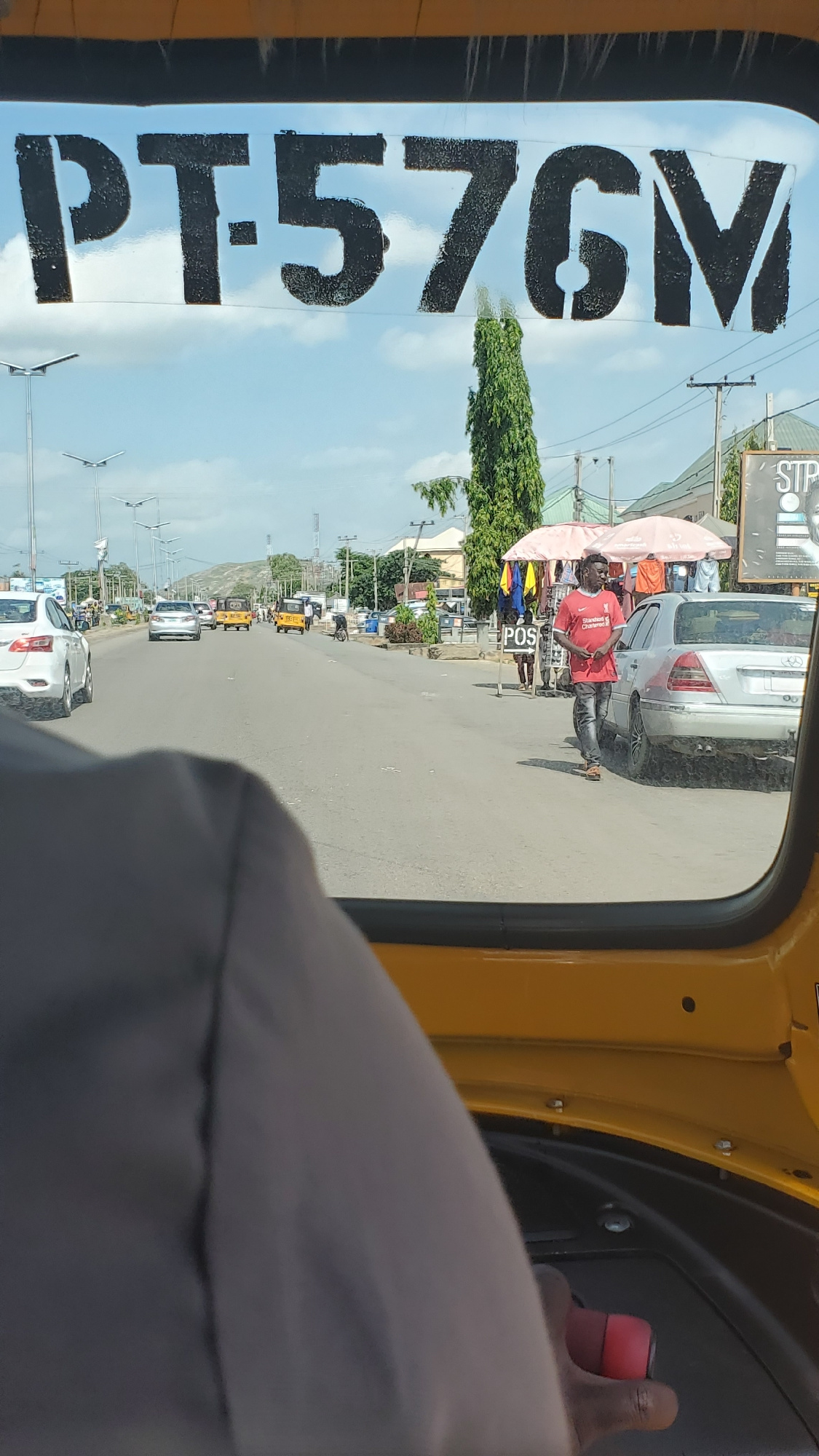 This media file captures a view from inside a yellow vehicle, likely a rickshaw or auto-rickshaw, traveling on a road in Jalingo, Nigeria. The perspective is from the driver's or passenger's seat, looking out through the side.
The main focus is the reflection on the side window, which shows a reversed image of the road and surroundings. Prominently displayed in large, black, stencil-like lettering on the window is the text "PT:57GV".
Outside, the scene depicts a street with several vehicles, including cars and other auto-rickshaws. A man in a red t-shirt and black trousers is standing near a silver car, holding a pink umbrella and a red object, possibly a bag. Other people are visible in the background, some standing on the roadside. There are trees and buildings lining the street, and power lines are visible overhead, suggesting an urban or semi-urban setting.
The sky appears to be a light blue with some clouds, indicating it's daytime, likely late morning or early afternoon given the lighting. The overall atmosphere seems ordinary, depicting everyday street life. There are streetlights visible along the road, and what appear to be some telecommunication towers in the distance. The road surface is asphalt.