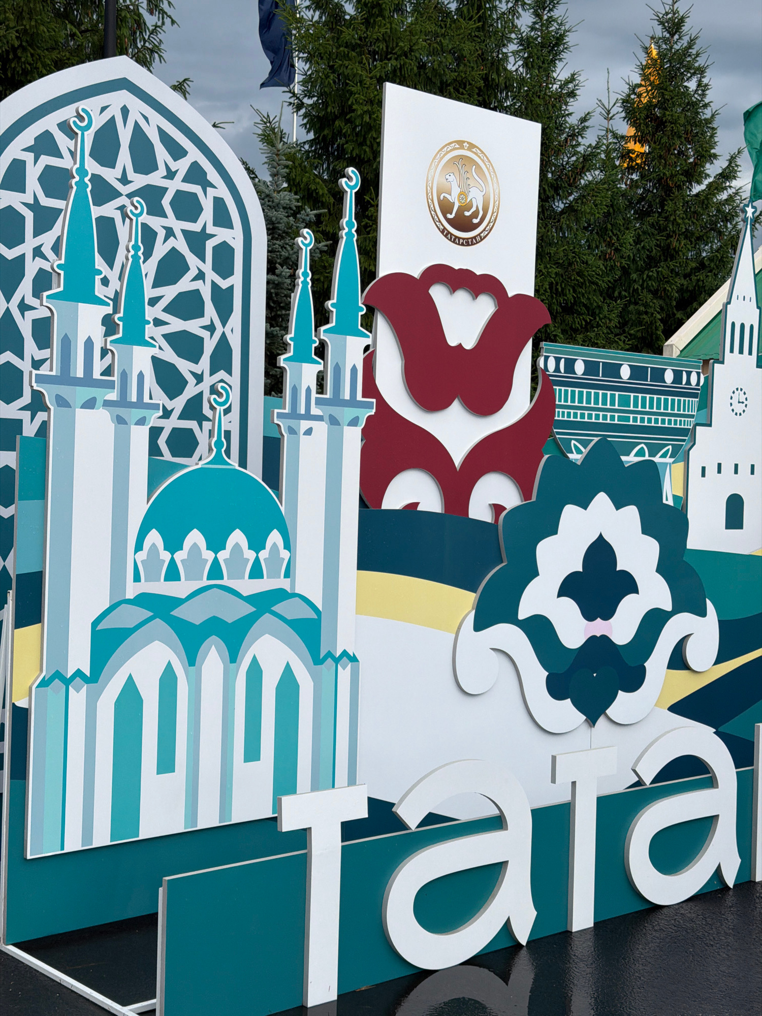 The image shows a colorful, stylized outdoor display featuring architectural elements reminiscent of Islamic and possibly Tatar culture, placed in front of evergreen trees under an overcast sky. Dominating the foreground is a large, white cutout of the word "Tata" in a bold, sans-serif font, with the letters appearing to be three-dimensional and positioned on a dark teal base.
To the left, a prominent cutout depicts a mosque with a large turquoise dome and multiple minarets, framed by an intricate, white geometric lattice pattern. The mosque features stylized arches and vertical elements in shades of light blue and white.
Further to the right, other decorative elements are visible. A large, stylized maroon floral or petal-like shape is positioned behind the mosque and slightly to the right. Above this, a white rectangular panel displays a circular emblem with gold detailing, featuring a mythical creature and text that appears to be in Cyrillic script, reading "ТАТАРСТАН" (Tatarstan). To the right of this panel, a cutout of a building with a clock tower and green roof is visible, resembling a church or historical structure. A dark teal band with a white horizontal patterned design runs across the middle ground. A large, dark teal and white floral motif with radiating petals is positioned to the right of the mosque and below the clock tower building. The entire display is set against a backdrop of dense, dark green evergreen trees and a gray, cloudy sky. The ground in front of the display appears wet, suggesting recent rain. There are no people visible in the image.