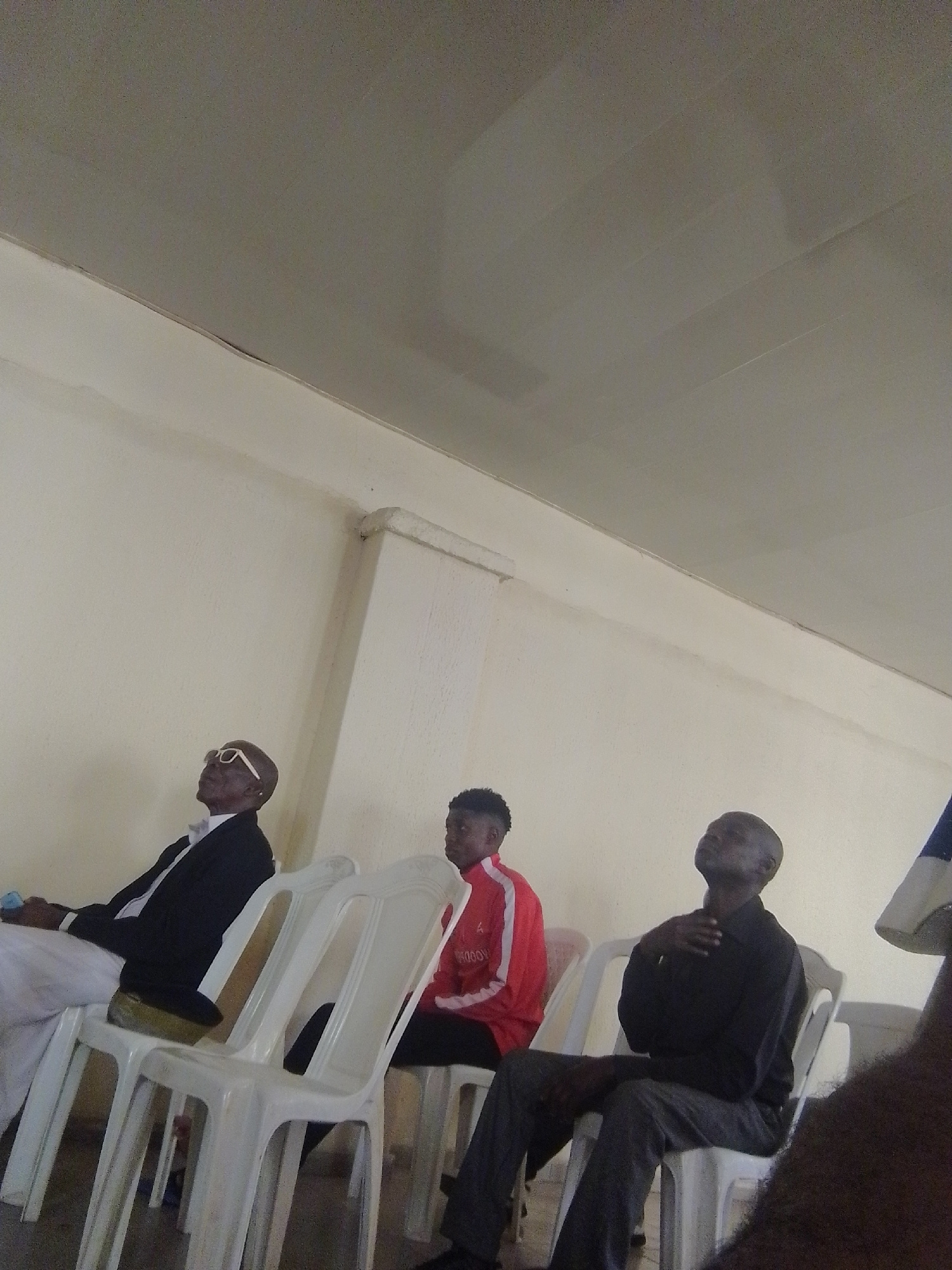 A low-angle, slightly blurry, medium shot shows three men seated on white plastic chairs against a pale yellow wall. The ceiling above is a textured, light gray. On the far left, a man in a black suit, white shirt, and bow tie, with white-rimmed glasses, looks upwards with his head tilted back. Next to him, a man in a red jacket with white trim, likely a tracksuit, also looks upwards. The third man, on the right, wears a black long-sleeved shirt and gray pants and looks upwards with his hand on his chest. The lighting is neutral and appears to be from overhead. The overall impression is of individuals attending an indoor event or service.