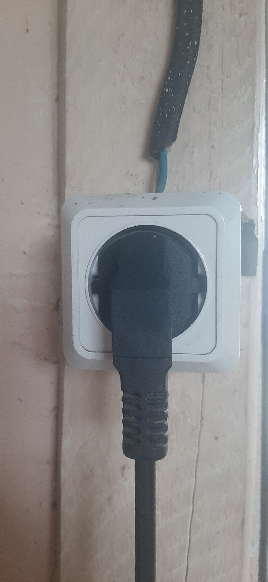 A black electrical plug is inserted into a white wall socket. The socket is square with rounded corners and has a circular opening. Above the socket, a black electrical cord with white speckles is taped to the wall, with a blue wire visible near the socket. The wall appears to be textured and painted a light color, with some scuff marks and a vertical crack. The image is a close-up, centered on the electrical outlet and plug. The location is identified as Yessentukskaya, Russia.