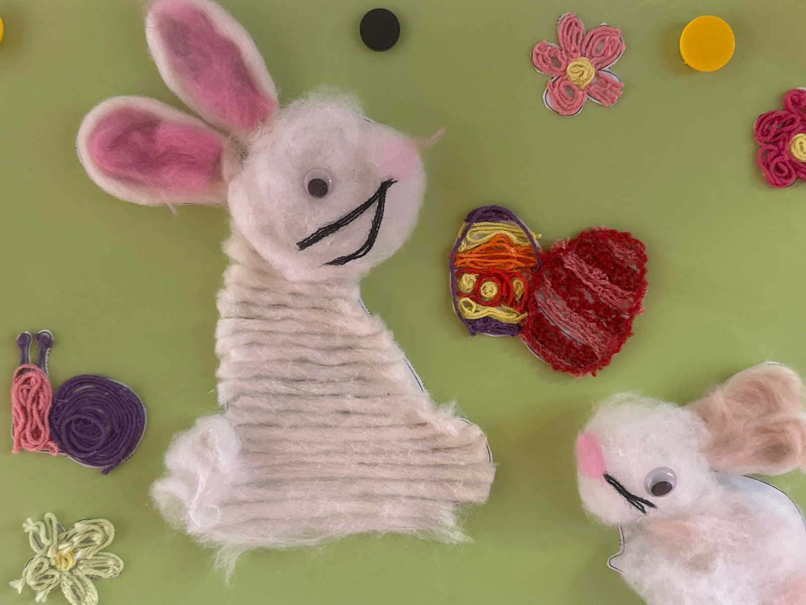 This is a flat lay, top-down, medium shot of a light green background adorned with various handmade Easter-themed crafts. In the center, a large white yarn-wrapped bunny figure is positioned on the left, facing right. Its body is covered in horizontal lines of white yarn, and it has pink yarn inside its long ears and pink felt cheeks. Its eyes are googly, and it has a stitched black smile. To the right of the large bunny is a small, similarly crafted white bunny, facing left, with pink yarn in its ears and pink felt cheeks, also with googly eyes and a stitched mouth.
To the left of the large bunny is a craft snail made of pink and purple yarn, with two purple antennae. Below the snail is a small, light green yarn flower.
Above and to the right of the large bunny, there are two yarn flowers, one pink and one darker pink, each with yellow centers. Between the flowers and above the bunnies are two circular yellow and black decorations, possibly buttons or painted circles. To the right of the large bunny and below the yarn flowers are two additional crafts: an Easter egg made of colorful yarn (purple, yellow, orange) and a heart shape made of red yarn.
The overall scene is static and appears to be a collection of craft projects arranged on a surface. There is no indication of a specific location within a city, as the background is a plain, unadorned color.