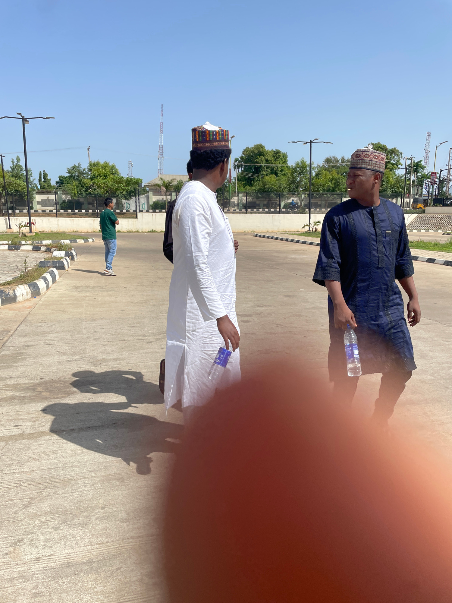 In this image, two men are standing outdoors on a paved surface, possibly a sidewalk or road, with a clear blue sky overhead, suggesting it is daytime and likely sunny. The man in the foreground is wearing traditional white attire and a patterned cap, and he is holding a bottle of water. The second man is dressed in a dark blue tunic and also wears a patterned cap. He is holding another bottle of water. Both individuals appear to be looking in the same direction, though their specific expressions are not entirely clear.
In the background, there is a scattering of trees and some buildings, including what appears to be telecommunications towers. The ground is a light-colored concrete, with black and white striped curbing along the edge of the paved area. There are shadows cast on the ground, indicating a bright light source, consistent with a sunny day.
A third person is visible further in the background, wearing a green shirt and blue jeans, also standing outdoors. The overall scene suggests a casual outdoor gathering or moment, possibly in a public space in Unguwar Rimi, Nigeria, given the clothing styles and general environment. There is no visible text in the image.