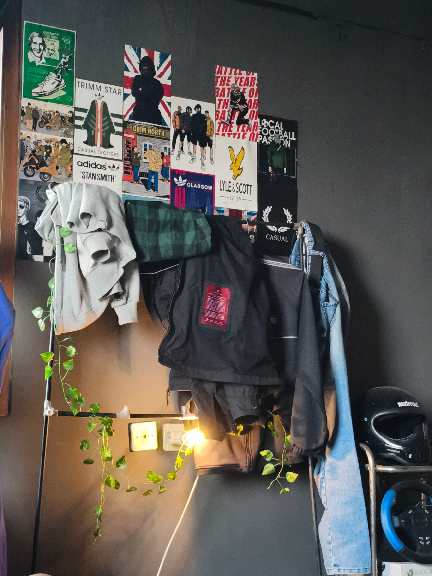 The image depicts a densely decorated wall featuring multiple posters with a casual, streetwear, and football fan aesthetic. The posters include brand logos like Adidas, Lyle & Scott, and graphics related to football and casual culture. Various clothing items are hung on a metal rack in front of the wall, including hoodies, a plaid shirt, a jacket, and jeans. A string of artificial ivy is draped over the rack and some of the clothing. There are also light fixtures and electrical outlets visible on the wall. On the right side of the image, a black motorcycle helmet and a blue-rimmed steering wheel are visible, suggesting a hobby or interest related to motorsports or gaming. The overall environment appears to be an indoor space, possibly a bedroom or a recreational area, characterized by its cluttered yet curated display. The city of Ciherang, Indonesia is not explicitly visible or suggested by any elements within the image.