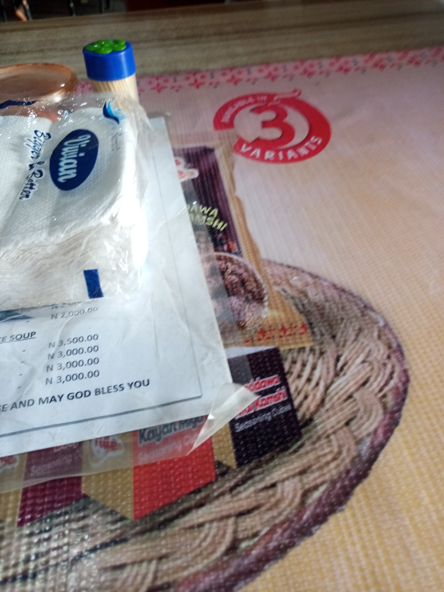 This media file captures a close-up, overhead view of a table surface with various items. The primary focus is on a stack of white, paper-like items, possibly napkins or food wrappers, with the brand name "Vivian" and the words "Bigger & Better" visible in blue text. The items appear to be sealed in plastic.
Beneath these, a white sheet of paper displays a list of prices in Nigerian Naira (N), with values such as N 3,500.00, N 3,000.00, and N 2,000.00. The phrase "AND MAY GOD BLESS YOU" is also printed at the bottom of this sheet.
To the right, a colorful, patterned placemat or tablecloth is visible, featuring a pattern of braided or woven textures in shades of brown and beige. A portion of a red circular graphic with the number "3" and the word "VARIANTS" in white text is also present on this surface.
In the background, a wooden surface can be seen, and a small blue object with green on top, possibly a toothpick holder, is also in the frame. Other items, including what appears to be a food container and some packaging with text like "AWA AMSHI" and "Seasoning Cubes," are scattered across the scene. The overall impression is that of a table set for a meal or a gathering, with various products and a receipt or order form. The lighting suggests an indoor setting, possibly during the day. No people are visible in the image.