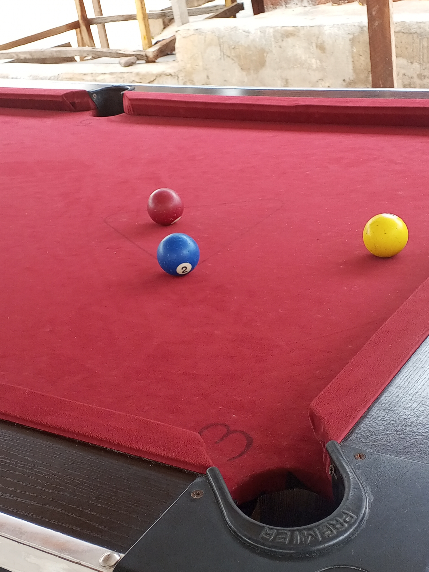 A close-up, eye-level shot shows three billiard balls on a red felt pool table. The balls are a solid red ball, a blue ball with the number "2" in white, and a solid yellow ball. The pool table has a dark wood veneer frame and a black pocket with the embossed word "PREMIER" on it. In the background, there is a concrete wall and some wooden structures, suggesting an outdoor or semi-outdoor setting. The lighting is bright, indicating it is daytime, and the weather appears to be clear. The scene is static, with no people present and no discernible activity other than the arrangement of the balls on the table. The background elements and the general appearance of the pool table suggest a casual, possibly recreational, environment. The location context indicates the scene is Lokoja, Nigeria.