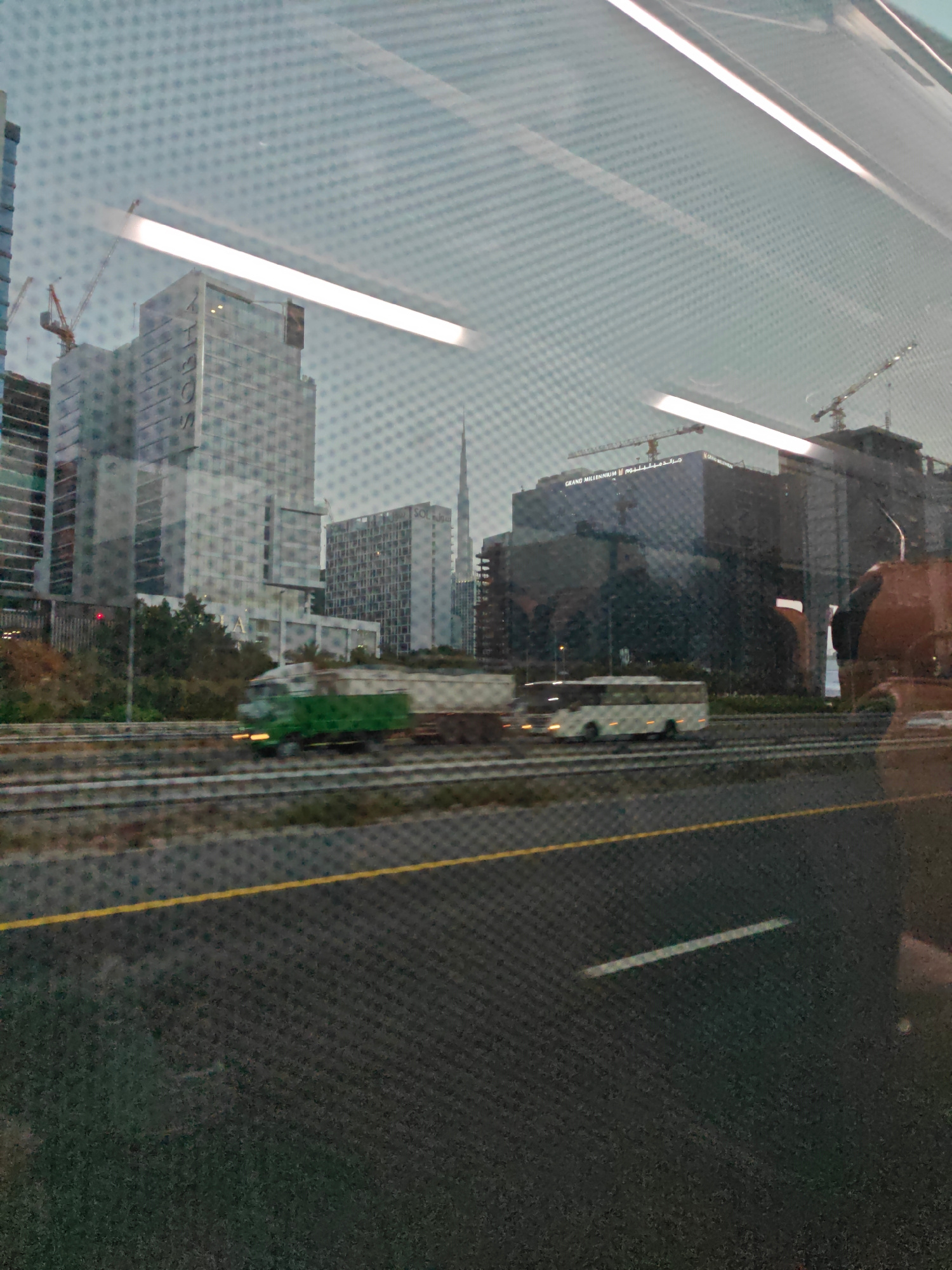 The image depicts a reflection on a surface, likely a window, showing a cityscape and a highway. In the background, several modern skyscrapers are visible, including buildings with the text "GRAND MILLENNIUM" and "SOFITEL". Cranes suggest ongoing construction. On the highway in the foreground, a green truck and a white bus are driving. The overall scene appears to be in Dubai, United Arab Emirates, given the prominent architecture and the presence of construction cranes. The reflection creates a grid-like overlay on the image.