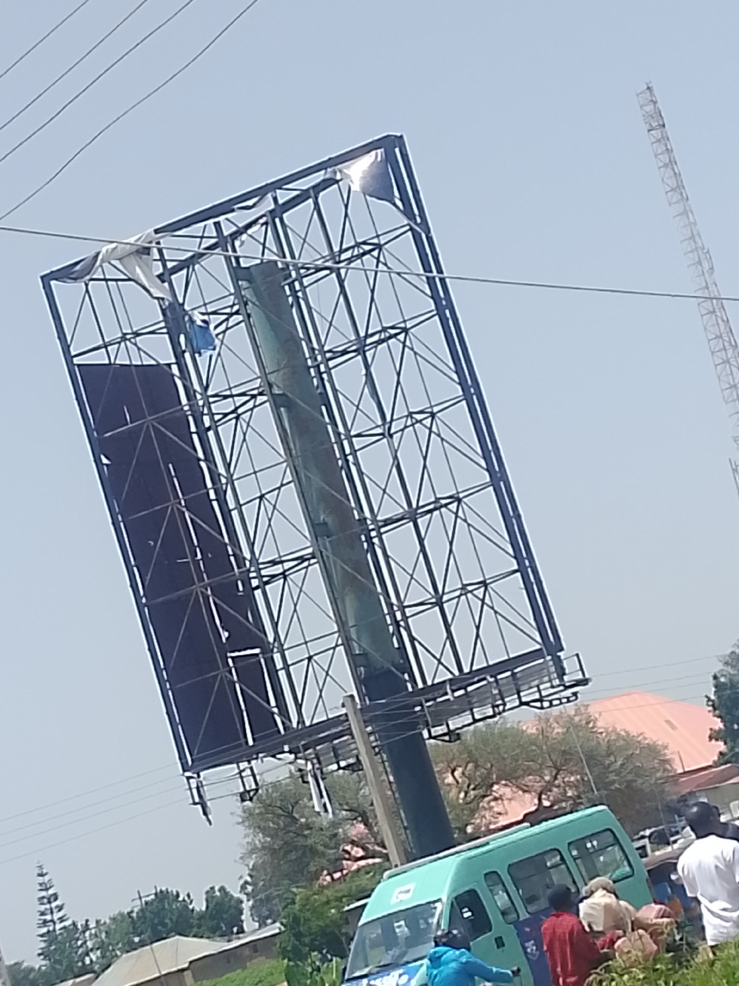A tilted billboard, made of dark metal framing, dominates the upper portion of the image. Some torn fabric, possibly from a previous advertisement, is caught on the frame. In the background, a tall, lattice-like tower stands against a pale sky, suggesting an outdoor, possibly rural or semi-urban setting.
Below the billboard, a teal-colored minibus is parked, with several people gathered around its open doors. Some individuals are wearing casual clothing. One person in a blue jacket is visible inside the bus, while others are seen interacting with those outside. The scene appears to be taking place during the daytime, with bright, diffused sunlight indicating a clear or slightly hazy sky. The presence of trees and some buildings in the lower background, along with the minibus, hints at a public space or thoroughfare in Bukuru, Nigeria. The overall impression is of a moment of everyday activity, possibly related to public transportation or people gathering around the bus. No visible text is discernable in the image.