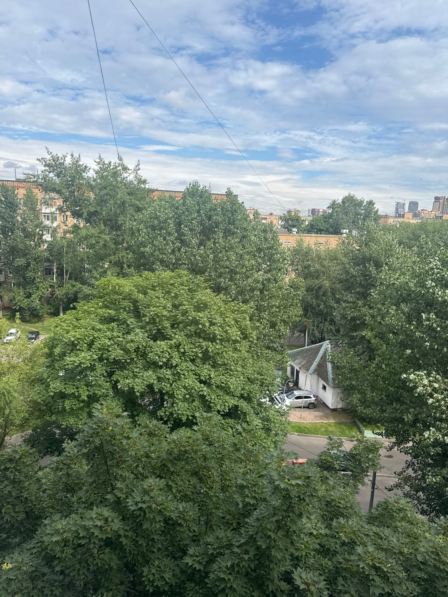 The image shows a view from a window overlooking a residential area in Moscow, Russia, on a cloudy day. In the foreground, dense green foliage from trees obscures the view. Behind the trees, there are several multi-story apartment buildings, typical of Soviet-era architecture. A small, white, gabled building with a corrugated roof is visible, with a silver car parked in front of it. Further in the background, more trees and buildings can be seen, with a distant view of modern high-rise buildings and a hazy skyline. Power lines are visible crisscrossing the sky. There are no people or animals visible in the image. The environment appears to be a mix of older and newer urban development, with abundant greenery.