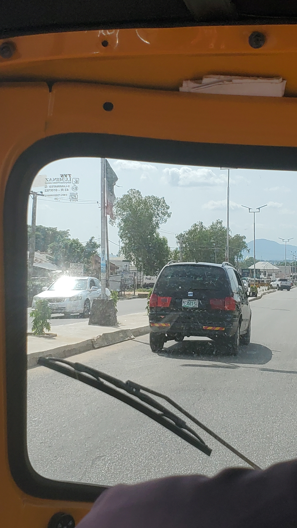 The image shows a view from inside a vehicle, looking out through the windshield and a side window. The vehicle appears to be yellow. Outside, there is a road with several other vehicles. The most prominent is a black car with a license plate reading "PE3521 SE". To the right of the black car, a white car is visible, reflecting bright sunlight. In the background, there are trees, buildings, and mountains under a bright, possibly sunny sky. The scene suggests a daytime setting, likely in an urban or semi-urban area. The presence of streetlights and the general road infrastructure indicate a populated location. A windshield wiper is visible in the foreground. The image is slightly tilted.