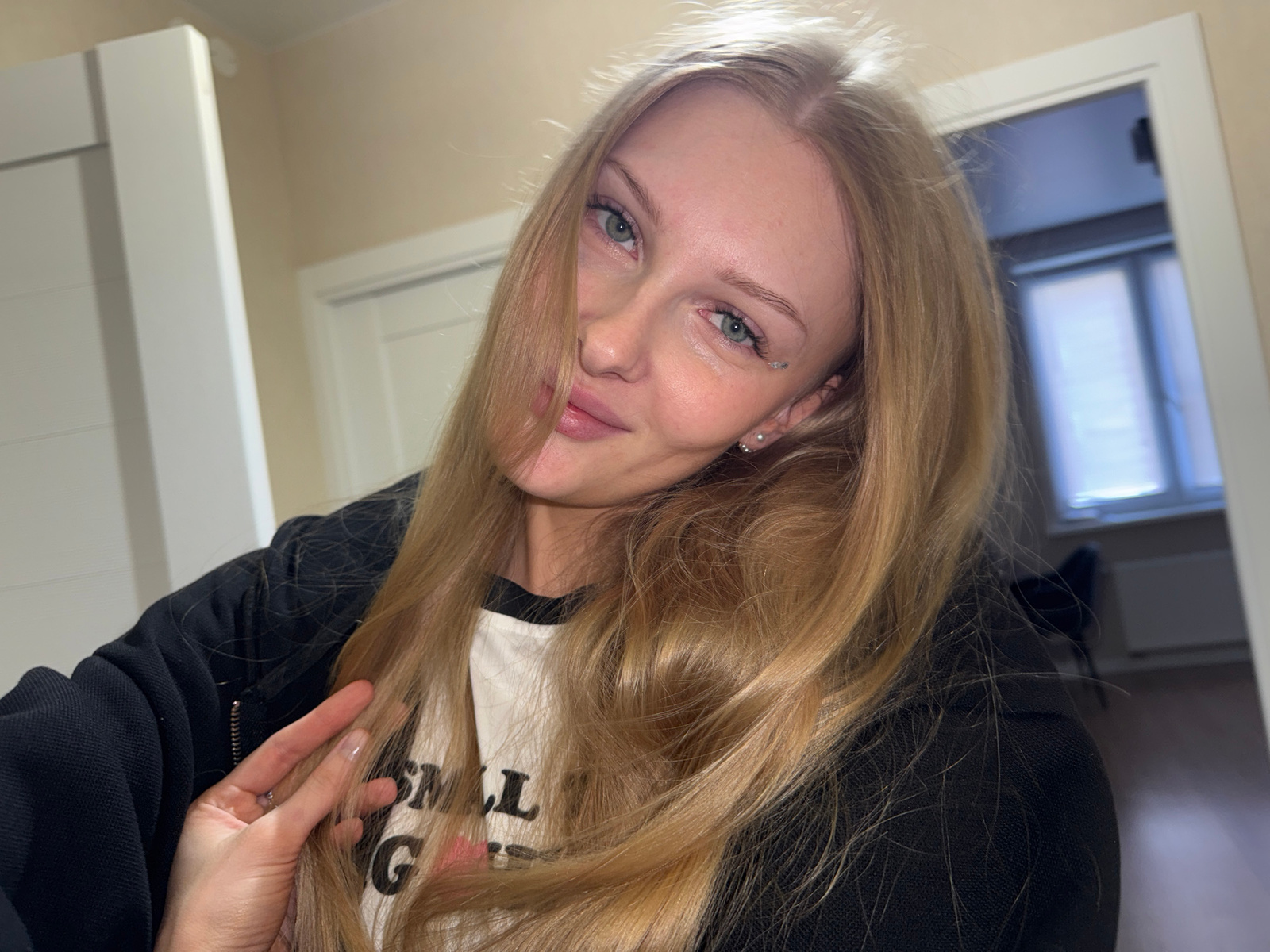 A young Caucasian woman with long blonde hair and blue eyes is taking a selfie in what appears to be a residential interior. She is wearing a black jacket over a white t-shirt with some text on it that is partially visible. Her hair is falling across her face, and she is holding a strand of it with her left hand. She has subtle makeup on, including eyeliner and possibly some glitter under her left eye. The background shows a doorway and part of a room with windows and a radiator. The lighting is soft, suggesting indoor lighting. There is no information in the image to indicate the specific location within the city of Perm, Russia.