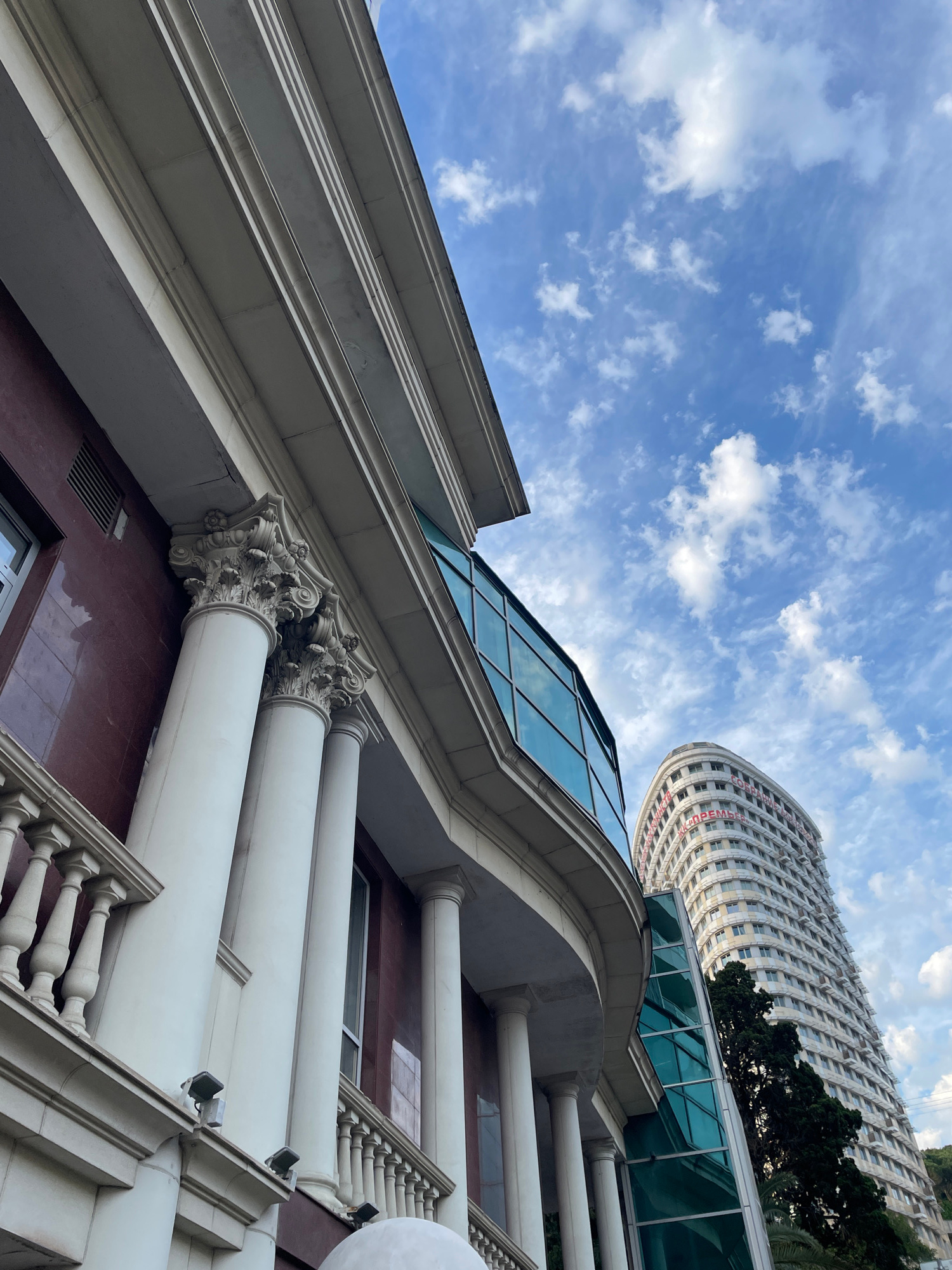 This low-angle shot captures an architectural scene under a bright, cloudy sky. The foreground features a building with a classical design, characterized by large white columns, ornate capitals, and a reddish-brown facade. A white balustrade with decorative spindles runs along the edge of a walkway or balcony. Above and to the left, a modern glass-paneled roof slopes upwards, reflecting the blue sky and scattered white clouds.
In the background, a distinctive, curved building with many windows rises into the sky. Red text is visible on the facade of this building. The overall setting suggests an urban environment, likely in Sochi, Russia, given the location context. The time of day appears to be daytime, with fair weather conditions indicated by the bright sky and fluffy clouds. The composition, with its upward perspective, emphasizes the grandeur and scale of the architecture. No people are visible in the frame, and no specific activity or event is taking place.