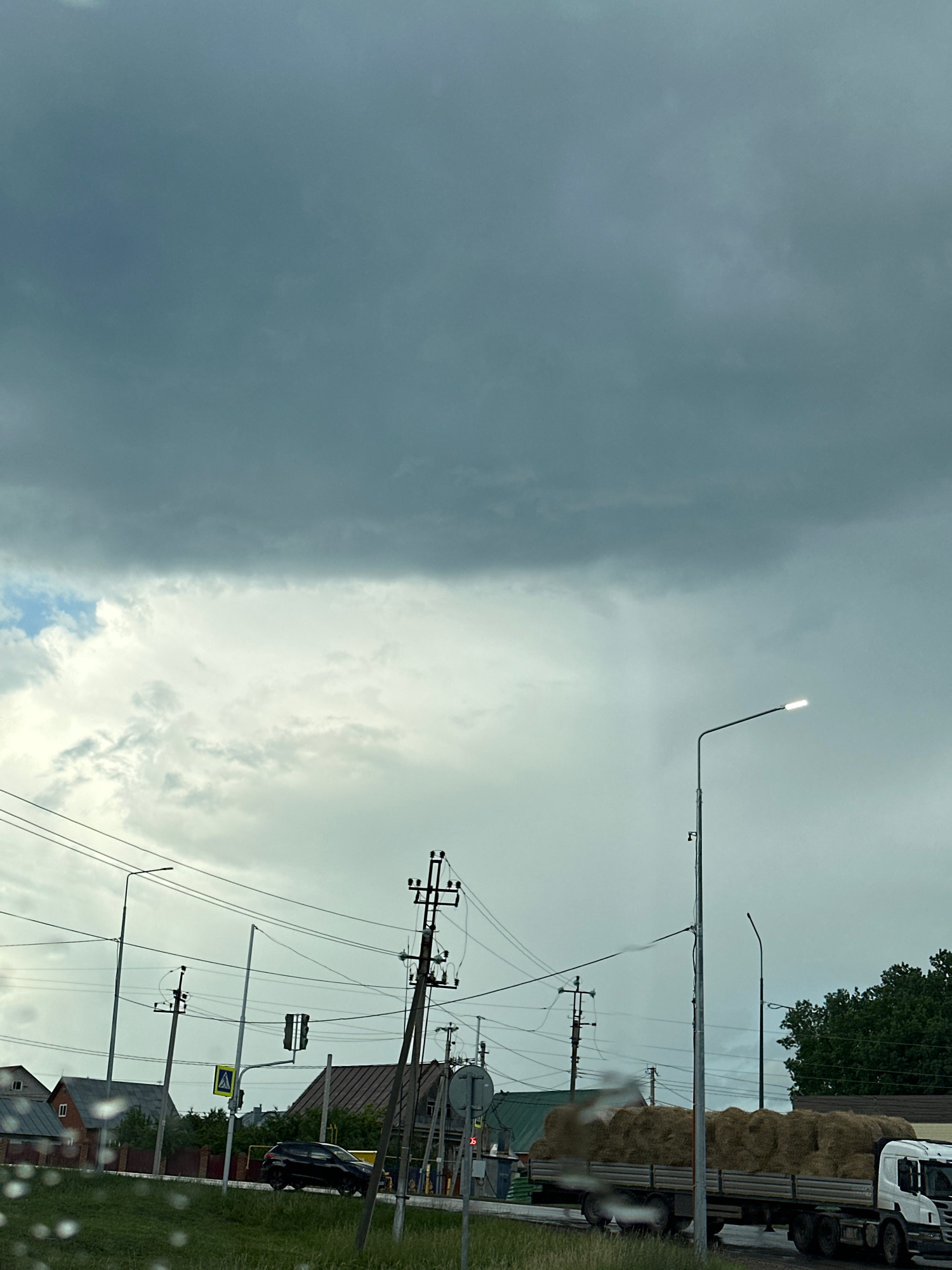 The image shows a split scene with dark storm clouds on the left and a brighter, partly cloudy sky on the right. A lone street lamp with its light on is visible in the brighter section of the sky.
In the right half of the image, a road is visible with a truck loaded with hay bales. There are utility poles with numerous wires, a traffic light, and a pedestrian crossing sign. A dark-colored car is parked on the side of the road. Buildings and trees are also present in the background.
The overall impression is of an impending storm. The weather appears overcast and possibly rainy, given the presence of water droplets on what appears to be the camera lens or a window. The lighting suggests it is daytime. The location cues, such as the architecture of the buildings and the road infrastructure, are consistent with a scene in Ufa, Russia. No people are clearly visible, and there is no discernible text in the image.