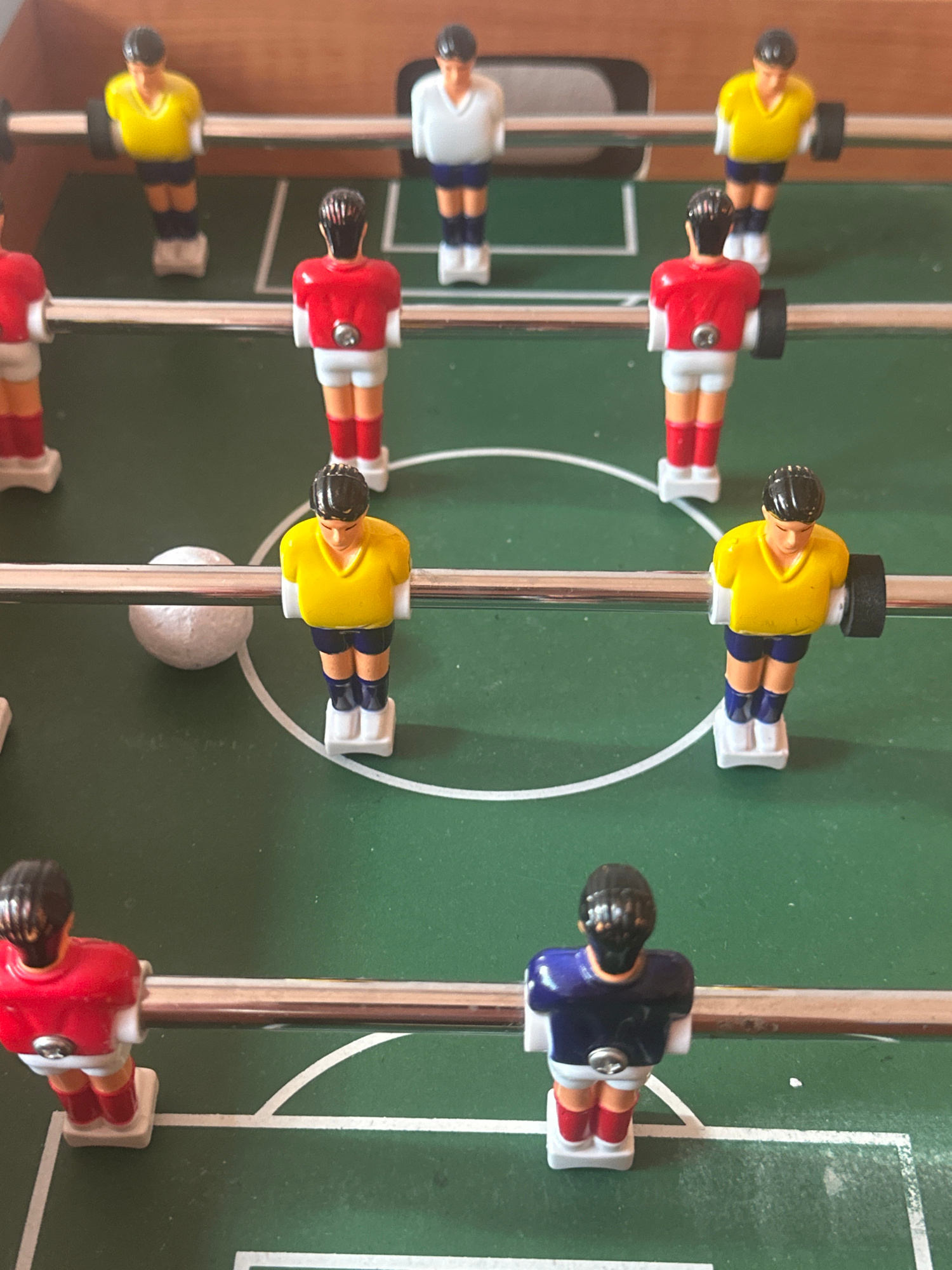 A tabletop foosball game is shown from a close-up, slightly elevated angle. The game board is green with white lines marking the field. A white foosball is positioned on the field, near the center. There are multiple miniature plastic figures representing soccer players arranged on metal rods that run horizontally across the table. The figures are wearing red, yellow, and blue jerseys with white shorts and shoes. Some figures have black hair. The background shows the wooden edge of the foosball table.