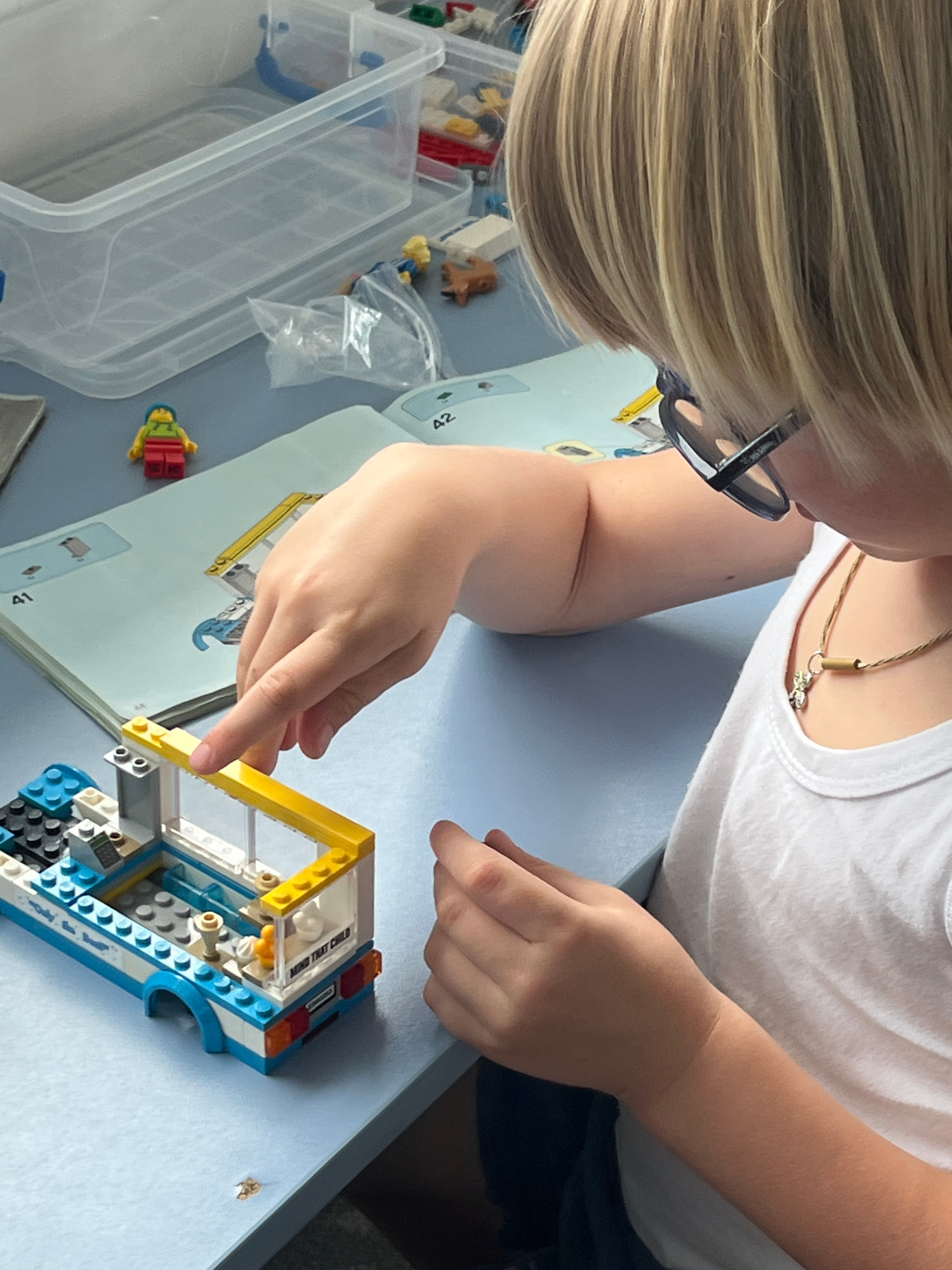 The image depicts a young blonde child wearing glasses and a white t-shirt, focused on assembling a LEGO vehicle on a light blue table. The child's hands are engaged with the LEGO bricks, specifically with a yellow and white structure. To the left, a LEGO minifigure with a yellow torso, red pants, and a blue hat stands on an instruction booklet, which shows diagrams and the number "42". A clear plastic storage bin is visible in the background, containing more LEGO pieces. The environment appears to be indoors, possibly a home or classroom setting, with natural lighting. There is no discernible information in the image that would suggest a location in the city of Yalta, Ukraine.