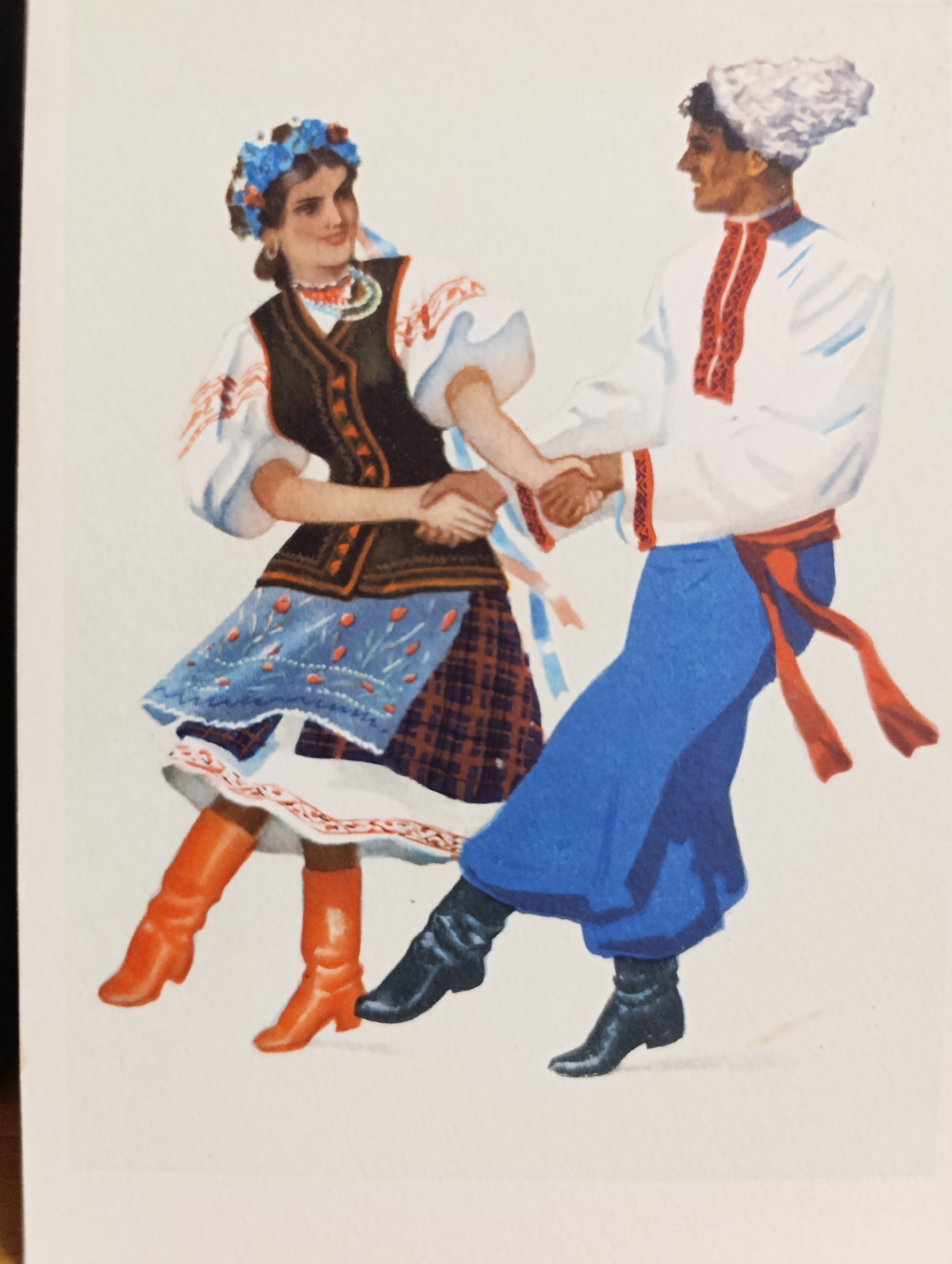 A full-body illustration depicts a man and a woman in traditional folk attire dancing. The woman is on the left, wearing a white blouse with red embroidery on the sleeves and collar, a dark vest over it, a blue embroidered apron, a plaid skirt, and orange boots. A blue floral headdress adorns her dark hair, and she wears a beaded necklace. The man is on the right, dressed in a white shirt with red embroidery down the front and on the collar, blue trousers, a red sash tied around his waist with dangling ends, and black boots. He wears a white, fluffy hat. They are holding hands and appear to be in motion, suggesting a dance. The background is a plain off-white color, offering no environmental context.