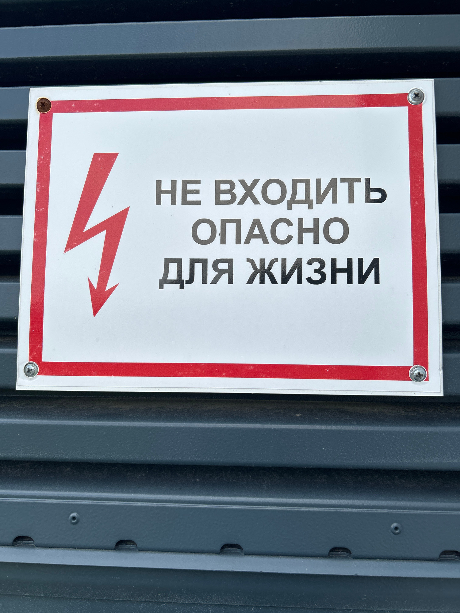 A white rectangular sign with a red border is affixed to a dark gray, corrugated metal surface. The sign features a prominent red lightning bolt symbol on the left side, pointing downwards. To the right of the symbol, in black Cyrillic text, are the words "НЕ ВХОДИТЬ" (Do Not Enter) on the top line, "ОПАСНО" (Dangerous) on the middle line, and "ДЛЯ ЖИЗНИ" (For Life) on the bottom line. The sign is secured to the metal surface by four silver screws, one in each corner. The overall impression is a warning sign, likely indicating an electrical hazard. The location is unknown.