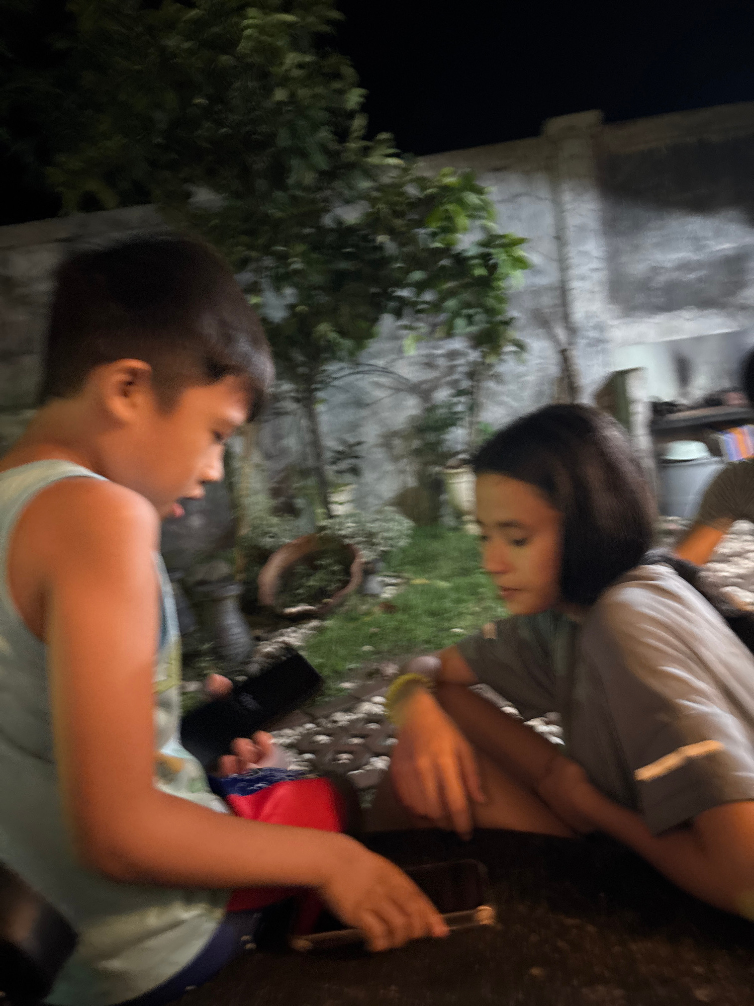 In Tagum City, Philippines, two young individuals are seated outdoors at night. The scene appears to be a backyard or patio area, with a concrete wall in the background and overgrown vegetation. The lighting is dim, suggesting it is evening or nighttime.
On the left, a young boy with short, dark hair, wearing a light blue tank top, is looking down at a black smartphone he is holding in his hands. A red and blue object, possibly a bag or a piece of clothing, is visible beside him.
To the right, a young girl with dark, shoulder-length hair, wearing a light grey t-shirt, is also looking downwards, her hands resting on her lap or a surface in front of her. She has a yellow bracelet on her left wrist.
The ground in front of them is a dark, textured surface, possibly a table. The overall atmosphere is casual and relaxed, with both individuals engrossed in their devices or simply resting. The background shows more greenery, with some potted plants or garden decor visible, and a rough, textured concrete wall.