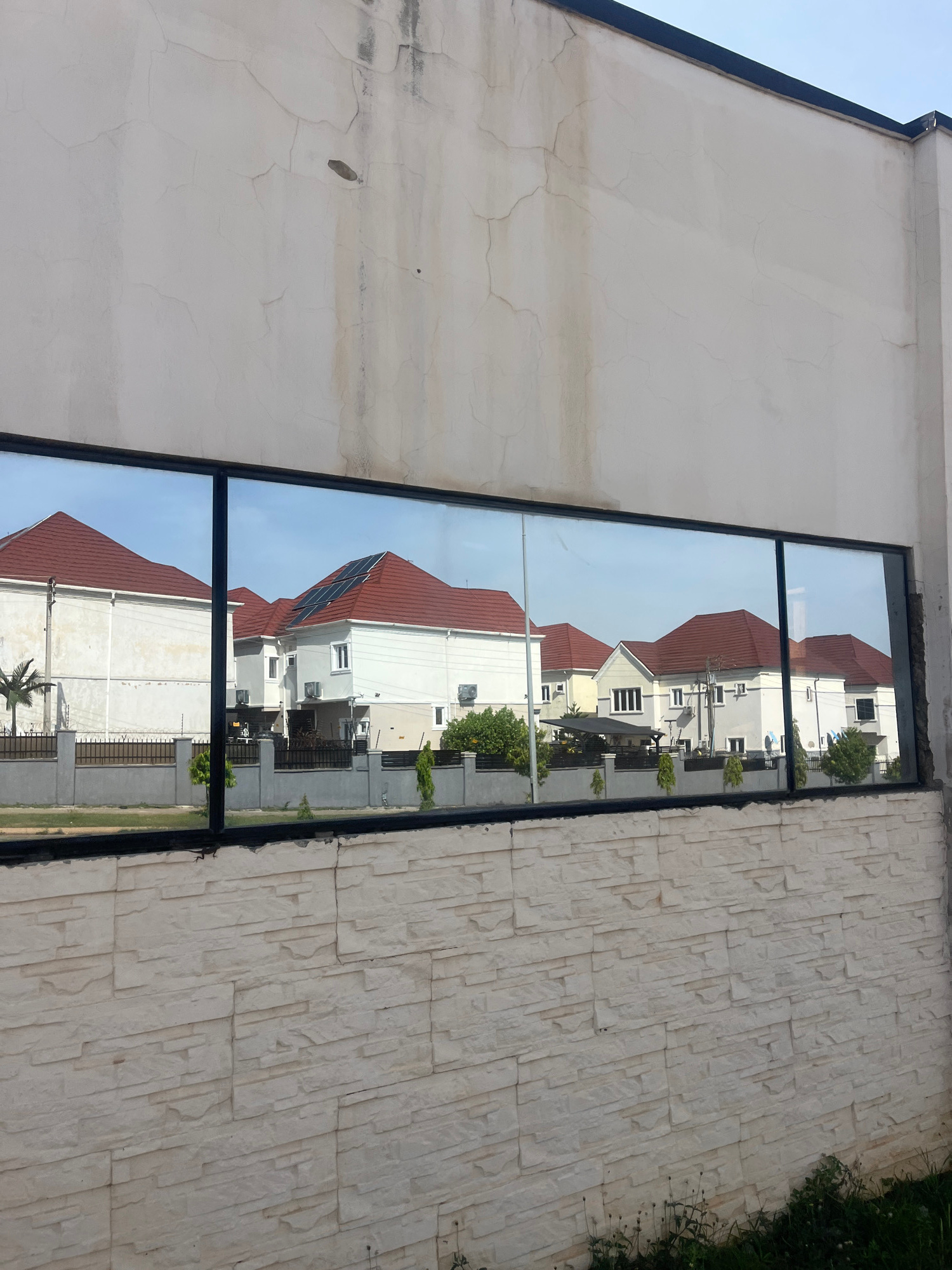 The image shows a building facade with a large, vertical window. The window reflects a row of houses with red tiled roofs and white walls. The houses appear to be modern, with multiple stories and several windows. Some of the houses have solar panels on their roofs. There are trees and greenery visible around the houses, suggesting a suburban or residential area.
The building facade itself is made of two different materials. The left side is a smooth, light-colored surface with visible cracks, indicating it might be plaster or stucco. The right side of the facade is covered in light-colored, textured stone tiles laid in a vertical pattern.
The window is divided into two sections by a horizontal mullion. The reflection in the window shows the houses from a slightly elevated perspective. The sky in the reflection is a clear blue, suggesting a sunny day. The overall scene depicts a residential area, likely in Karum, Nigeria, given the location context. There is no visible text in the image. The lighting suggests it is daytime, and the clear sky points to fair weather. The scene conveys a sense of quiet residential life.
