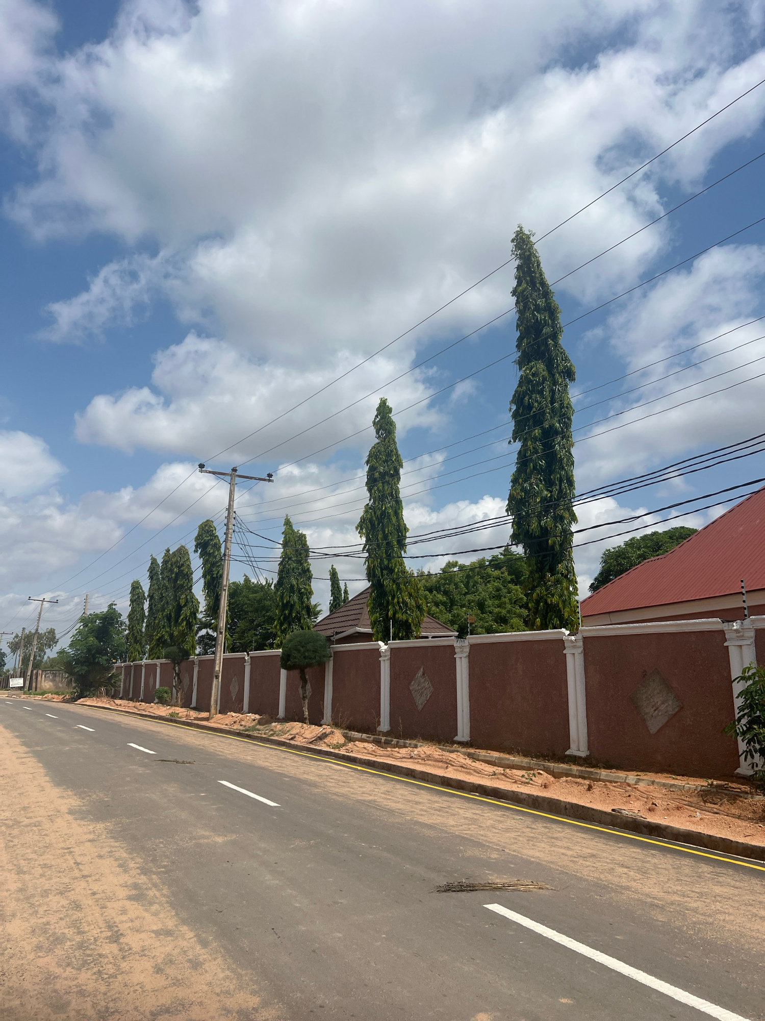 The image shows a paved road with white lane markings, bordered by a dusty shoulder. Along the right side of the road is a tall, reddish-brown wall with white decorative pillars and diamond-shaped accents. Behind the wall, several tall, slender trees with dark green foliage stand in a row, reaching towards a bright blue sky filled with fluffy white clouds. A red-roofed building is visible behind the trees and wall on the far right. Power lines are strung across the sky, supported by utility poles. The scene appears to be in a developed area, possibly a residential or commercial district within the city of Howayi, Nigeria. There are no people visible in the image.