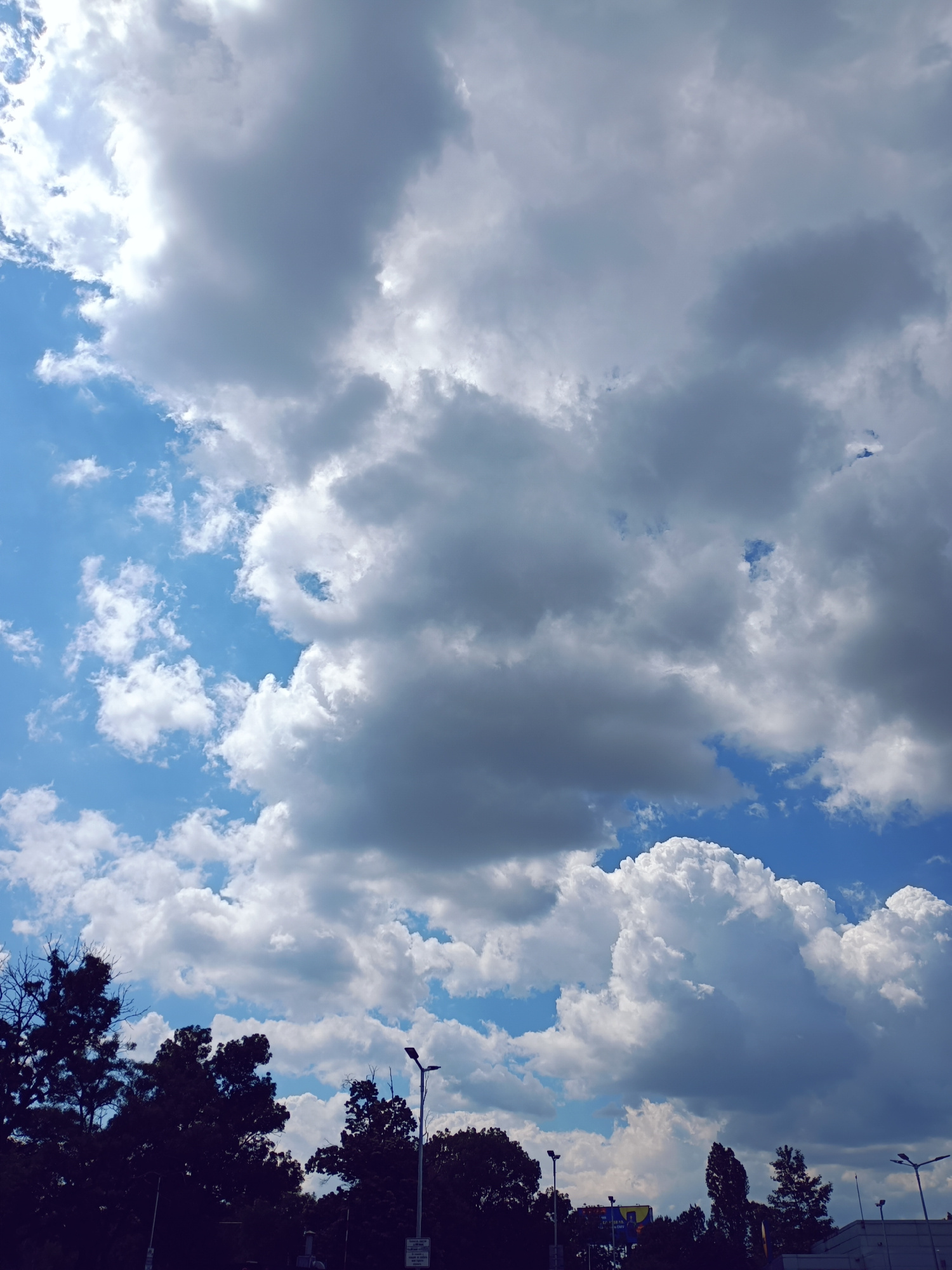 The image captures a sky filled with dynamic clouds, ranging from bright white to dark gray, against a vibrant blue backdrop. The sun is visible, casting a bright light on the upper left portion of the sky. Silhouetted against the sky are the tops of several trees and streetlights, suggesting an urban or suburban environment. In the distance, there are hints of buildings and possibly a billboard. The perspective is from a low angle, looking upwards. This scene is likely from Sofia, Bulgaria, given the typical urban and natural elements seen in the picture.