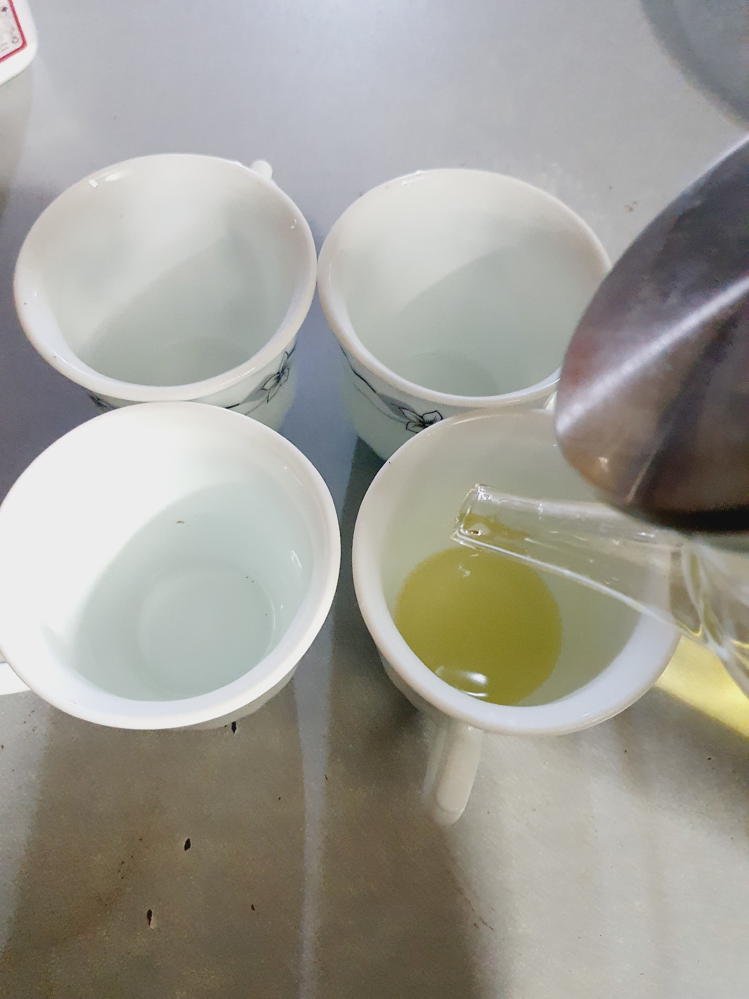 A top-down, close-up shot shows four white ceramic cups arranged in a rectangular formation on a mottled gray surface, possibly a countertop or table. Three of the cups are empty, revealing their pristine white interiors. The fourth cup, positioned in the lower right quadrant of the frame, contains a pale yellow liquid, and a stream of the same liquid is being poured into it from a clear glass teapot spout, which is partially visible entering the cup from the top right. The teapot itself is mostly out of frame, but its metallic lid is just visible at the very top edge of the image. The cups appear to be of a standard size for tea or coffee. The lighting is bright, suggesting an indoor setting. The image does not contain any discernible elements that would suggest a specific location within the city of Shiraz, Iran, or any particular activity beyond the pouring of a beverage.