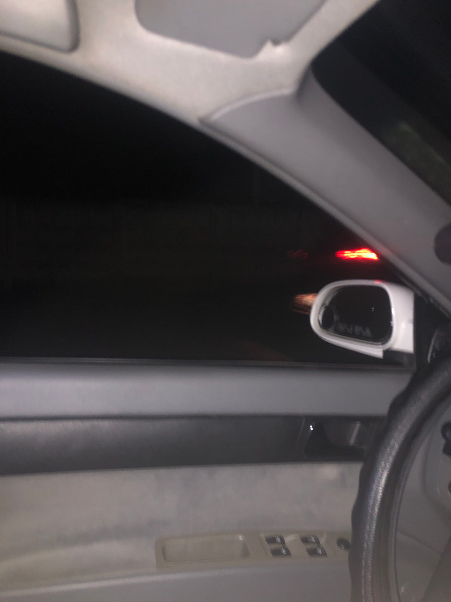 The image shows the interior of a car at night, looking out the driver's side window. The dark exterior is illuminated by the red taillights of another vehicle. A white side-view mirror is visible, reflecting the red lights and the interior of the car. The steering wheel is partially visible on the right side of the frame. The door panel and window controls are also visible. There are no people visible in the image. The scene appears to be on a road within the city of Khujakent, Uzbekistan.